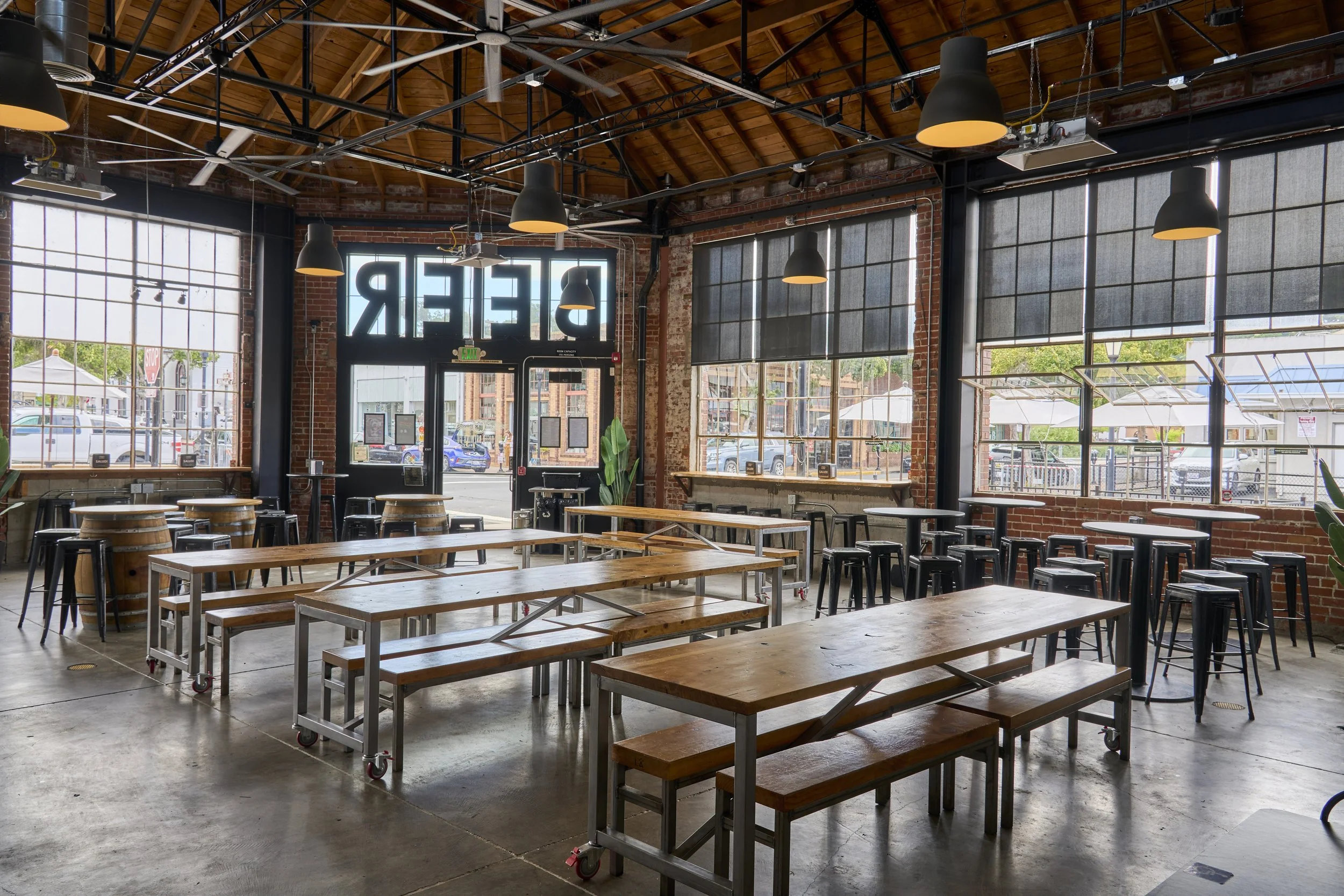 Interior of a modern warehouse-style restaurant with large windows, wooden tables, black stools, and a sign that reads 'BEER' inside.