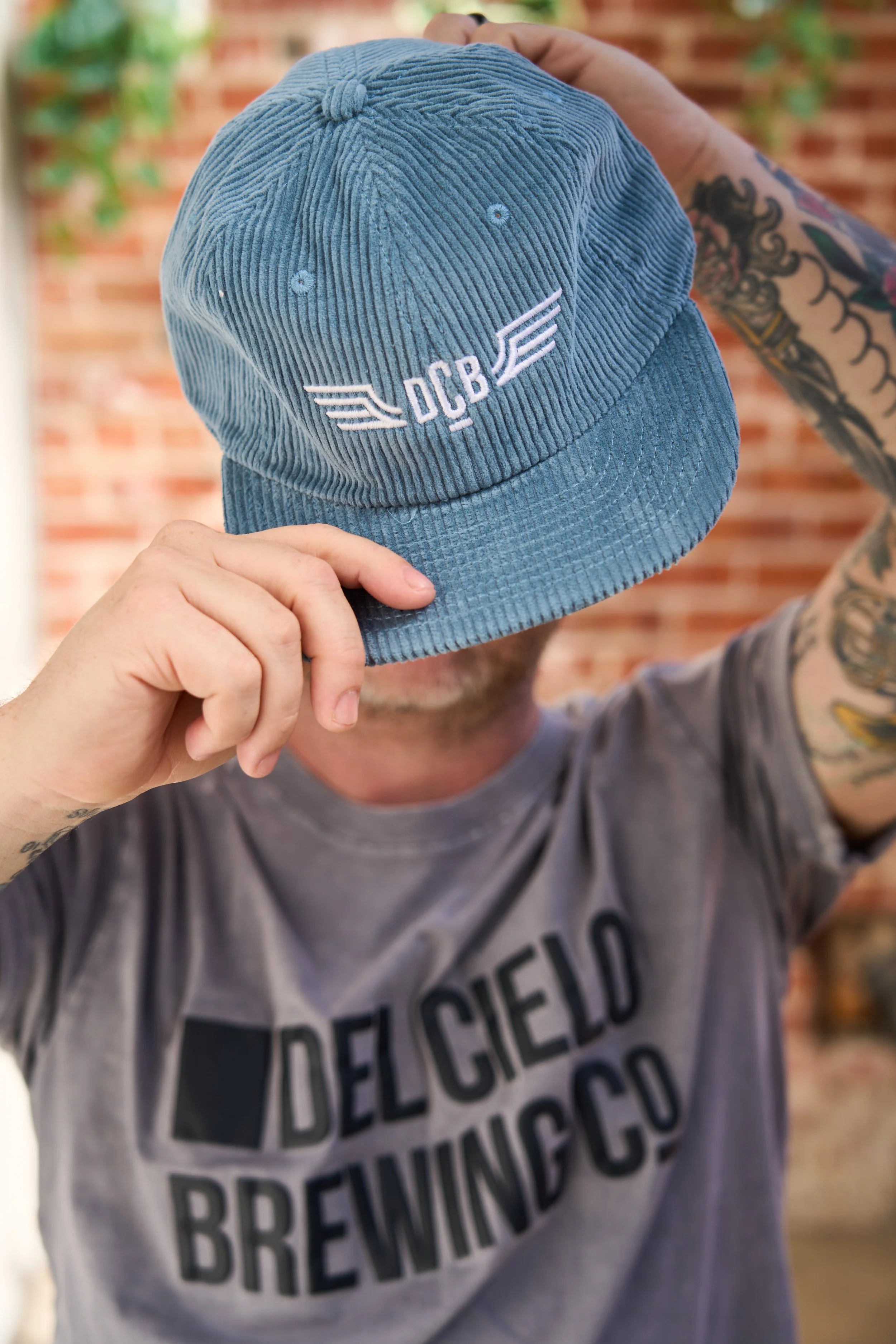 Person holding a blue corduroy cap with an embroidered logo reading 'DCB' and wing designs, tattoos visible on arm, standing in front of a brick wall and green plants.