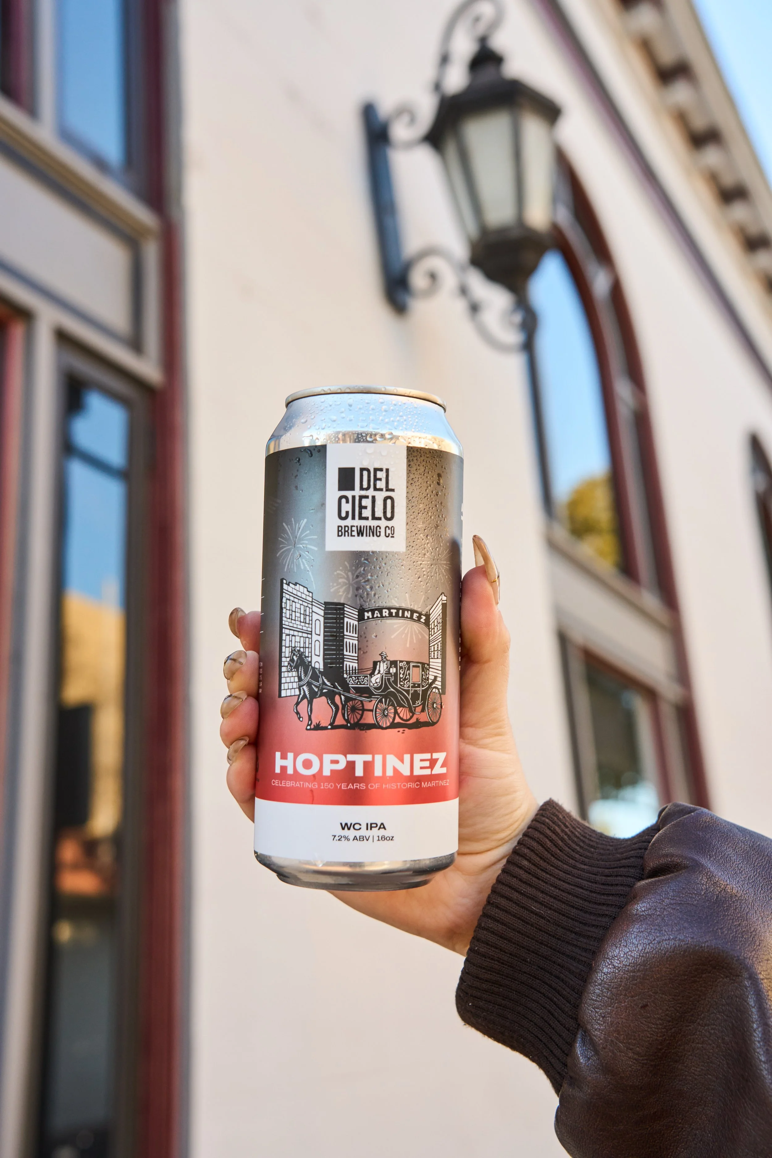 A hand holding a can of Hoptinez beer in front of a building with a black lantern-style light fixture and windows reflecting the sky.