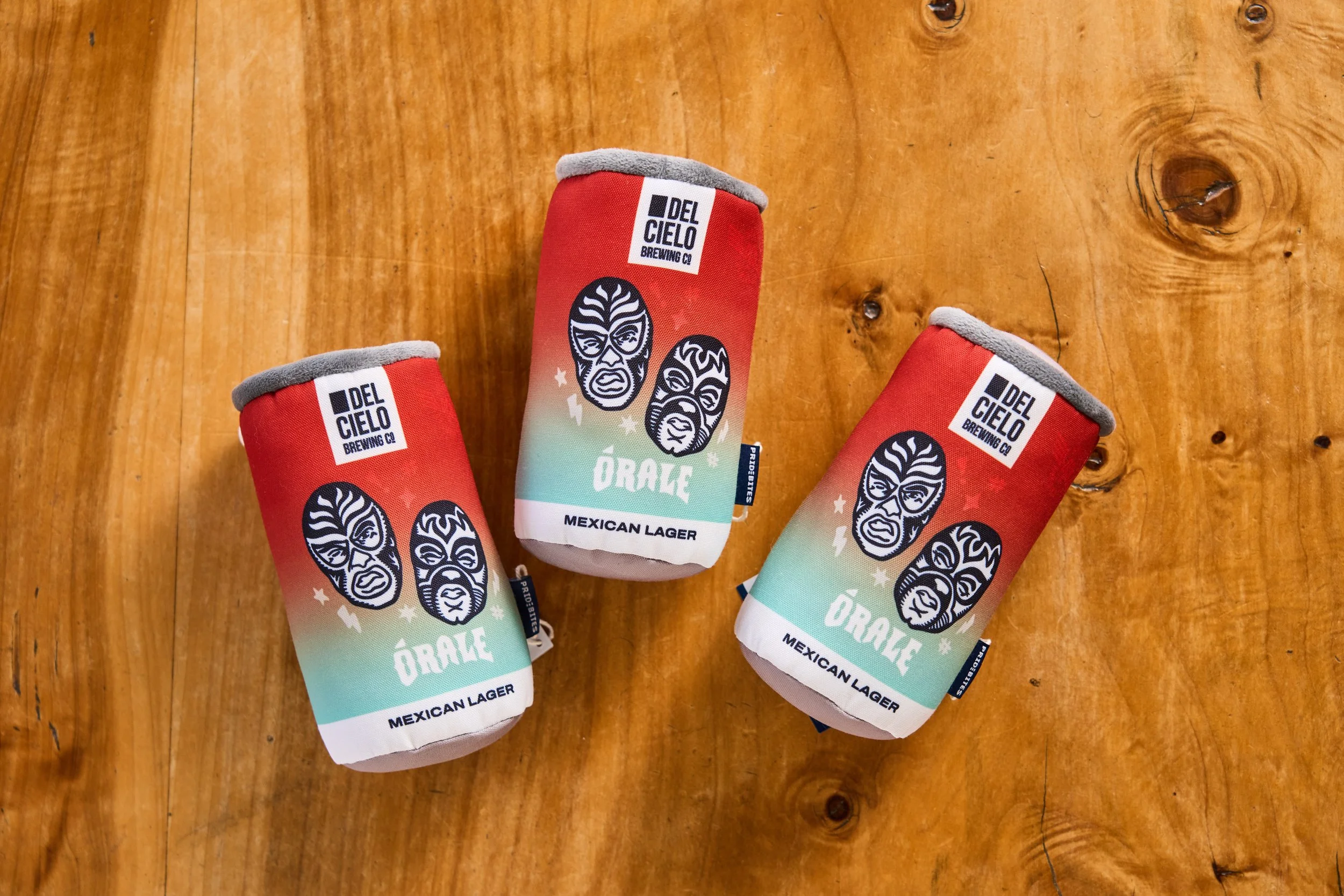 Three cans of Del Cielo Brewing Co. Mexican Lager beer placed on a wooden surface, with colorful labels featuring black and white faces and the word 'Orale'.