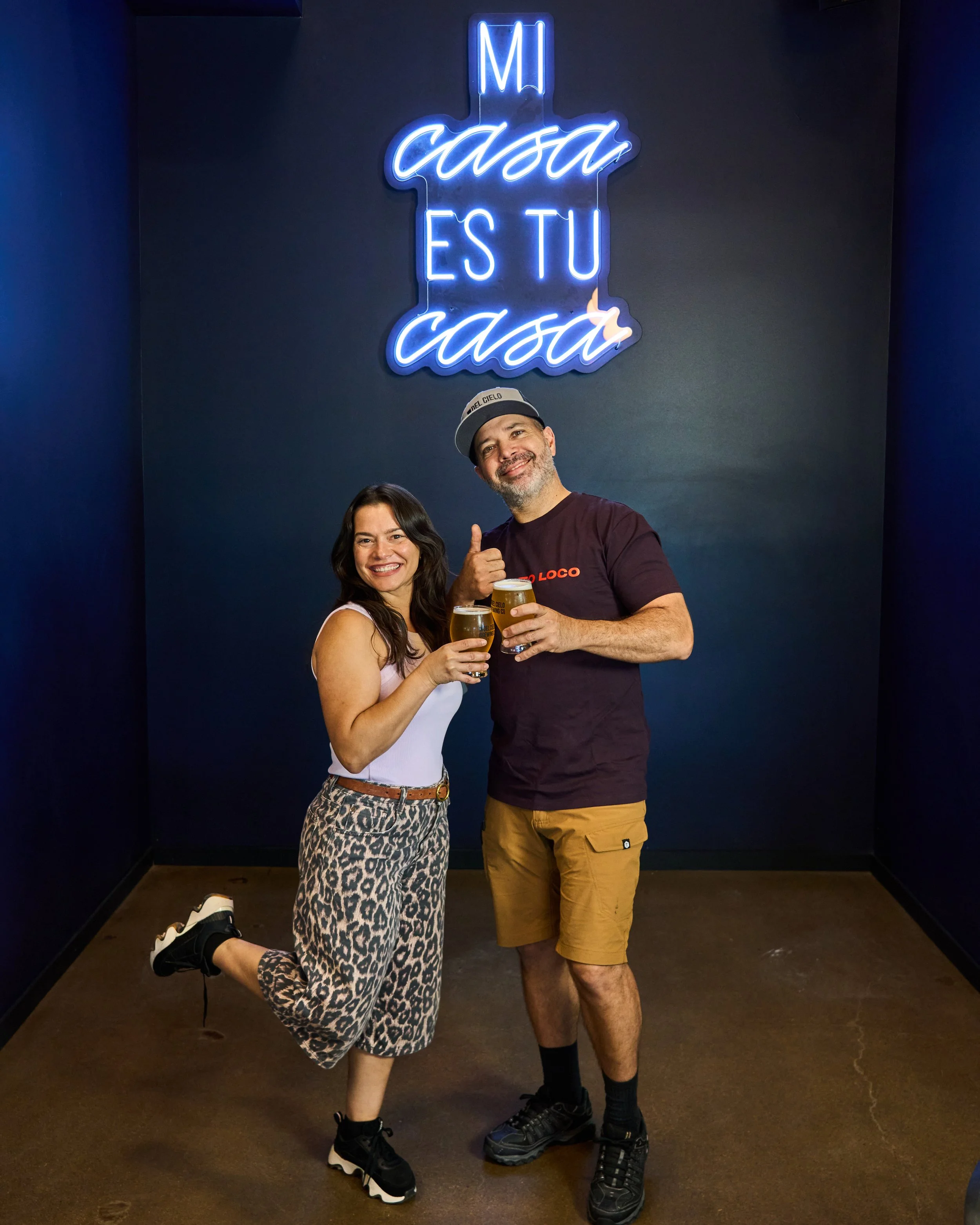 A man and woman standing and smiling in front of a dark wall with neon signs that read 'Mi casa es tu casa.' The woman is wearing leopard print pants, a white tank top, and black sneakers, and is kneeling on one knee. The man is wearing khaki shorts,