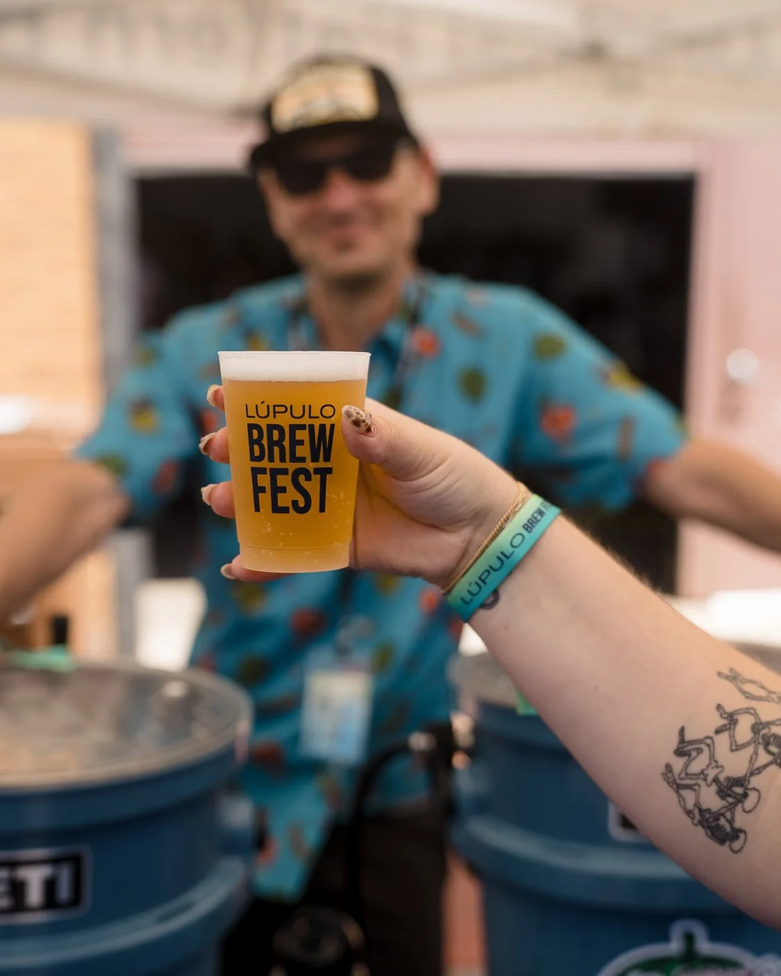 Person holding a plastic cup of beer with 'Lúpulo Brew Fest' printed on it, at a beer festival, with a smiling man in sunglasses and a colorful shirt in the background.