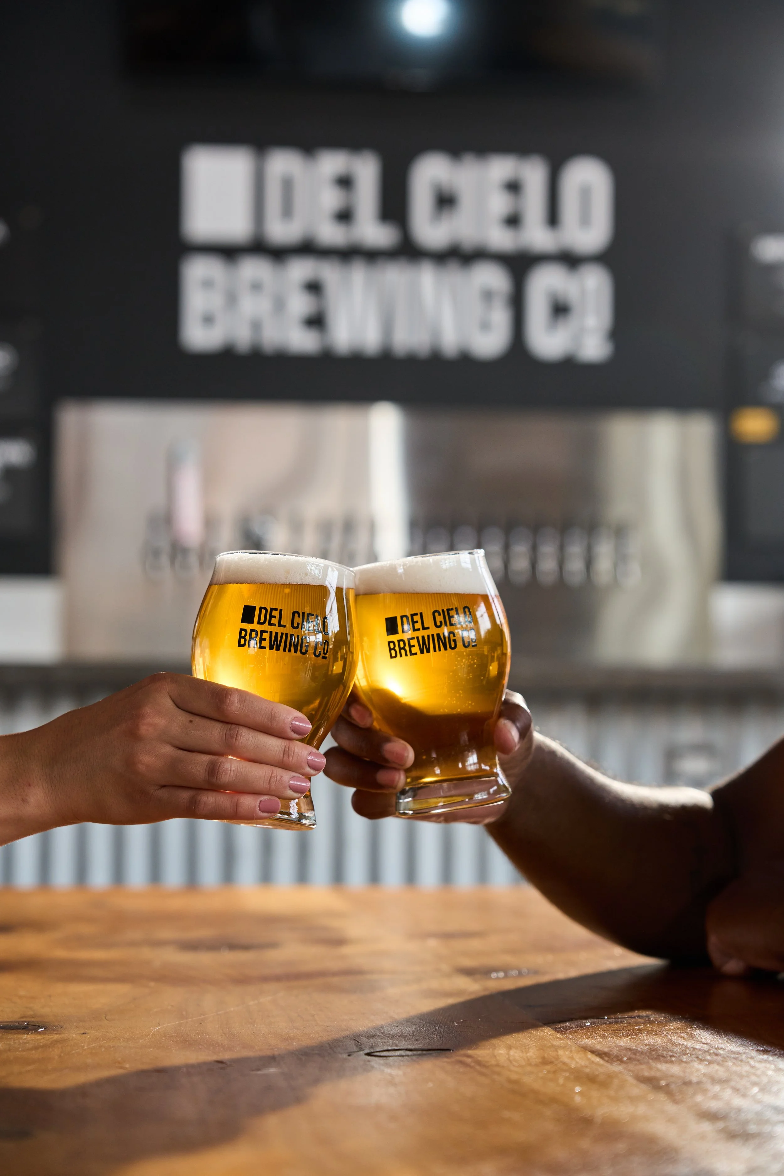 Two hands clinking glasses of beer at a bar, with the logo of Del Cielo Brewing Co on each glass, in front of a brewery background.