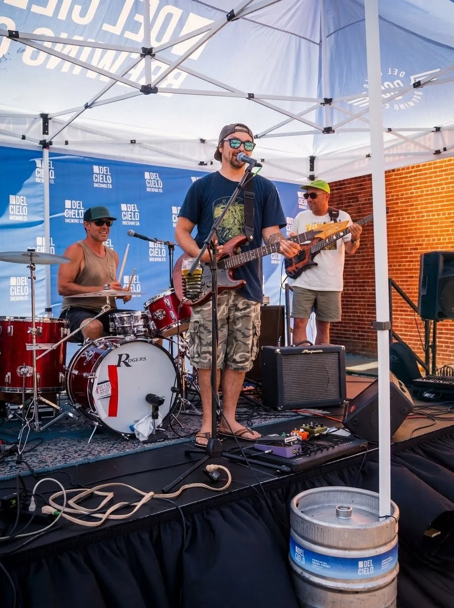 A young band performing on an outdoor stage under a white canopy with blue banners behind them. The band includes a drummer, a guitarist/singer, and a bass guitarist. The drummer is wearing sunglasses and a sleeveless shirt. The singer/guitarist is wearing sunglasses, a hat backwards, and camouflage shorts. The bass guitarist is wearing a hat, sunglasses, a white t-shirt, and shorts. There is a beer keg on the ground in front of the stage with a blue and white label.