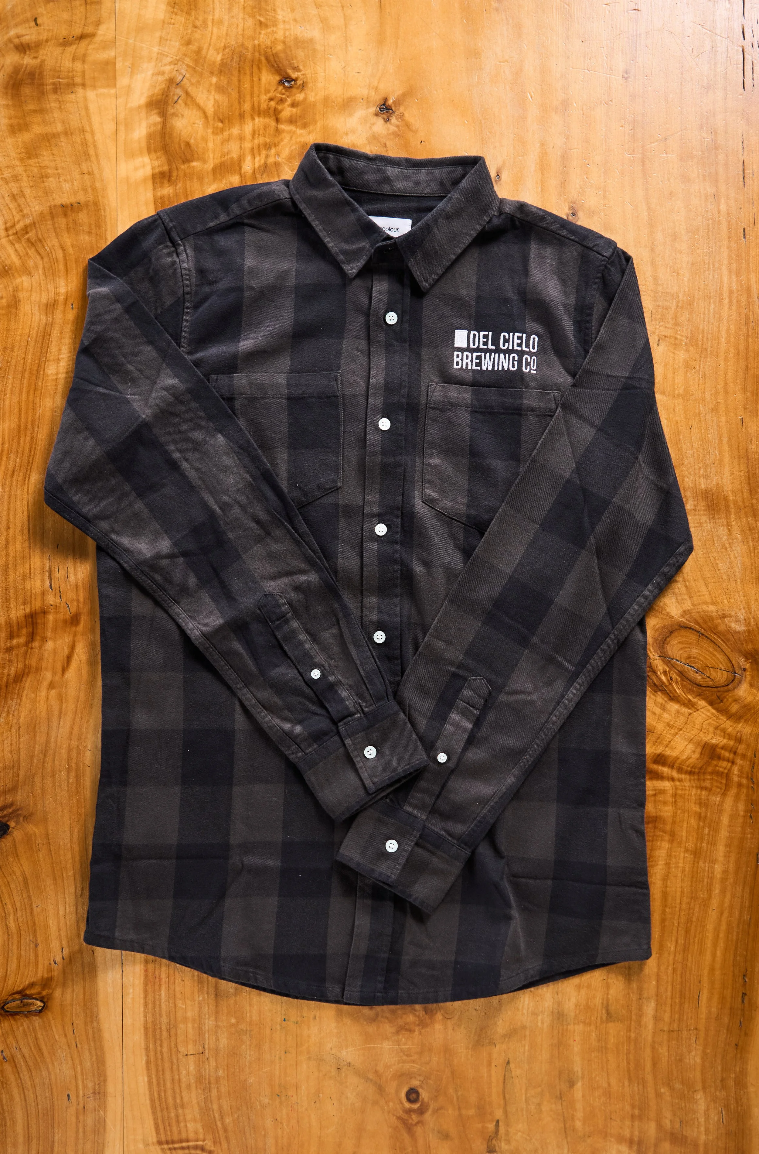 Black plaid button-up shirt with embroidery on the chest that reads 'Del Cielo Brewing Co,' laid on a wooden surface.