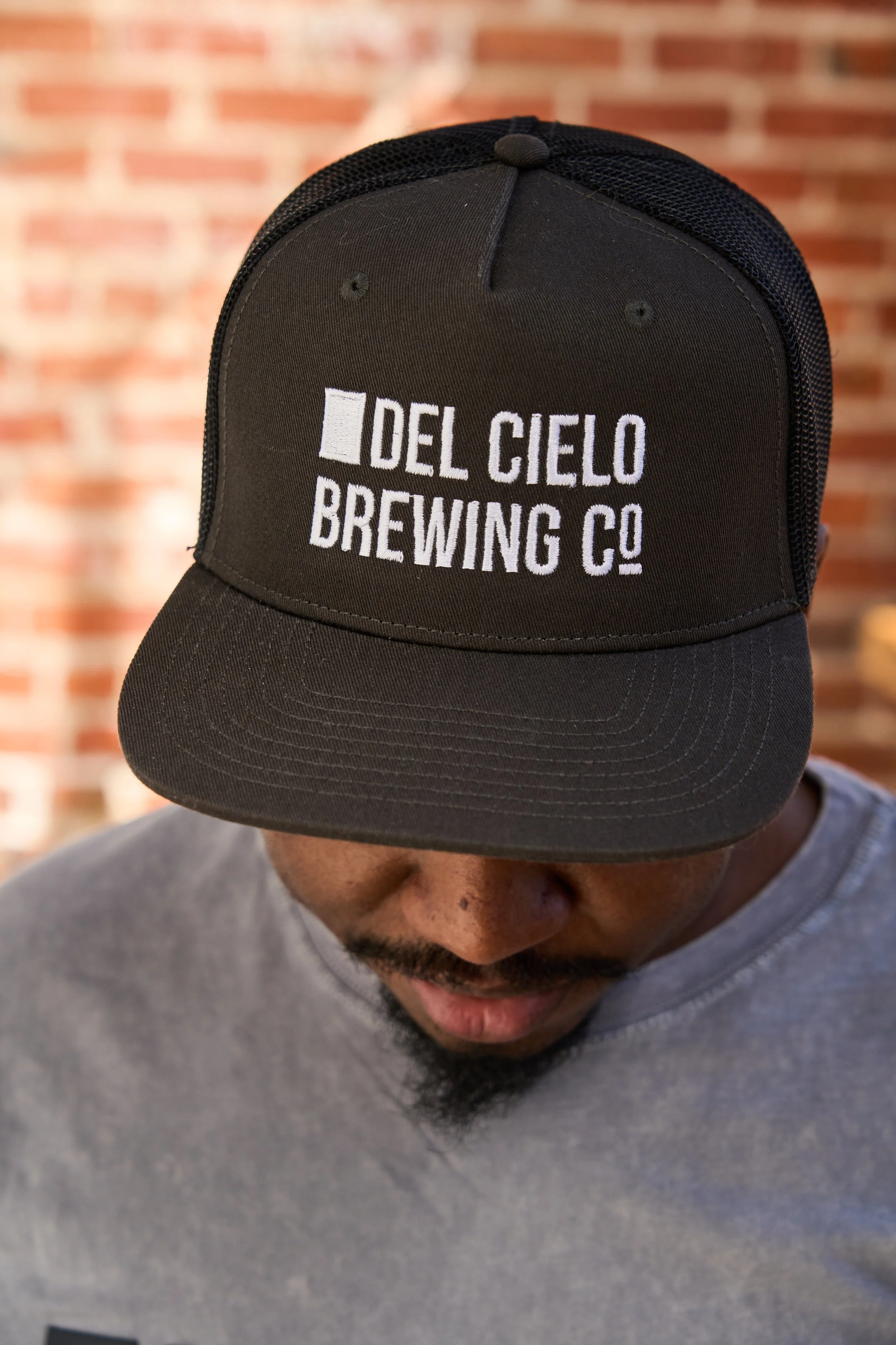 A man wearing a black trucker hat with 'Del Cielo Brewing Co.' embroidered in white, with a brick wall background.