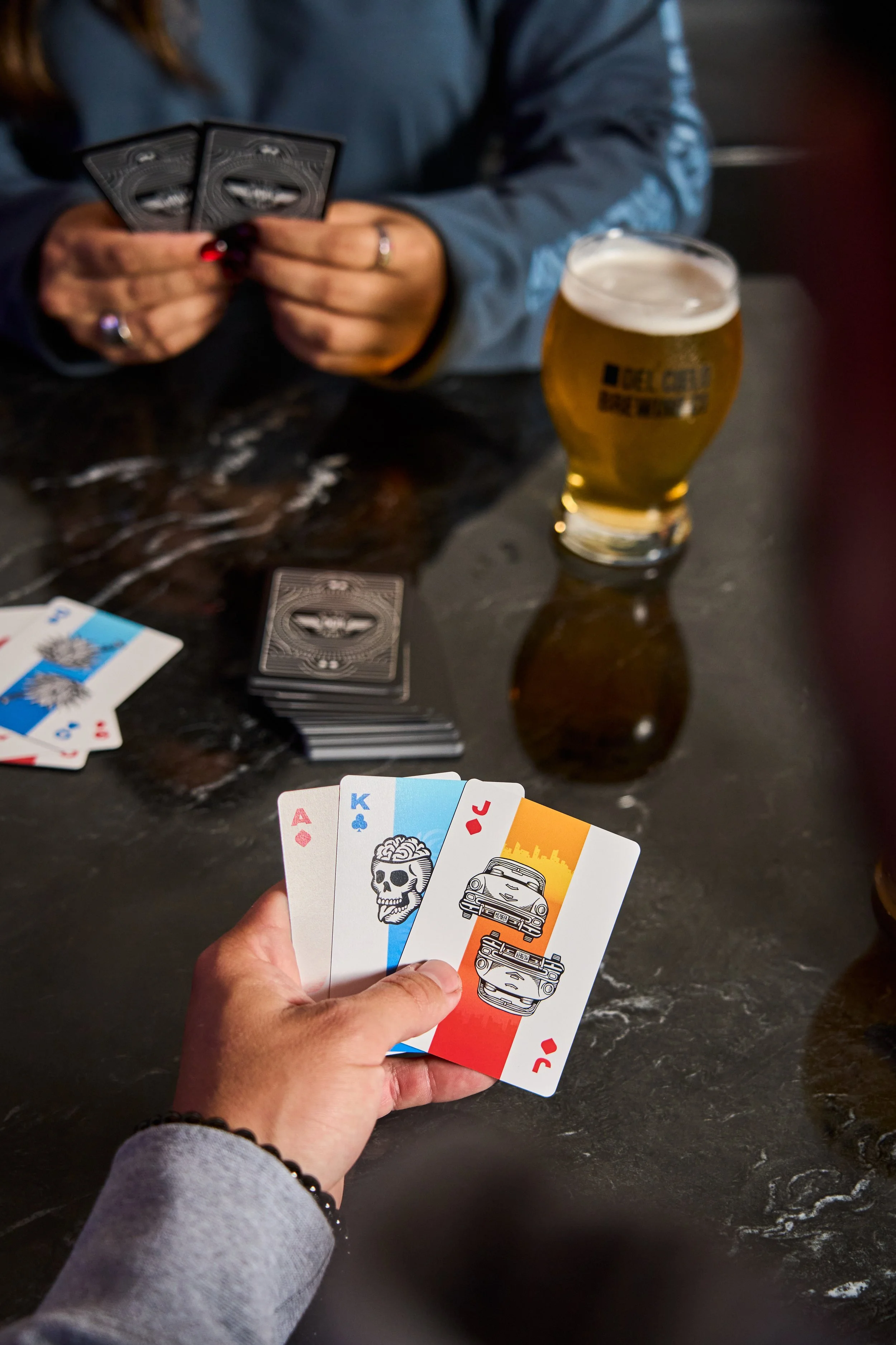 A person wearing a gray long sleeve shirt holding four illustrated cards, featuring a skull, a car, a cityscape, and a king of clubs. In the background, a table with a deck of cards, a glass of beer, and some playing cards with the Ace of diamonds an