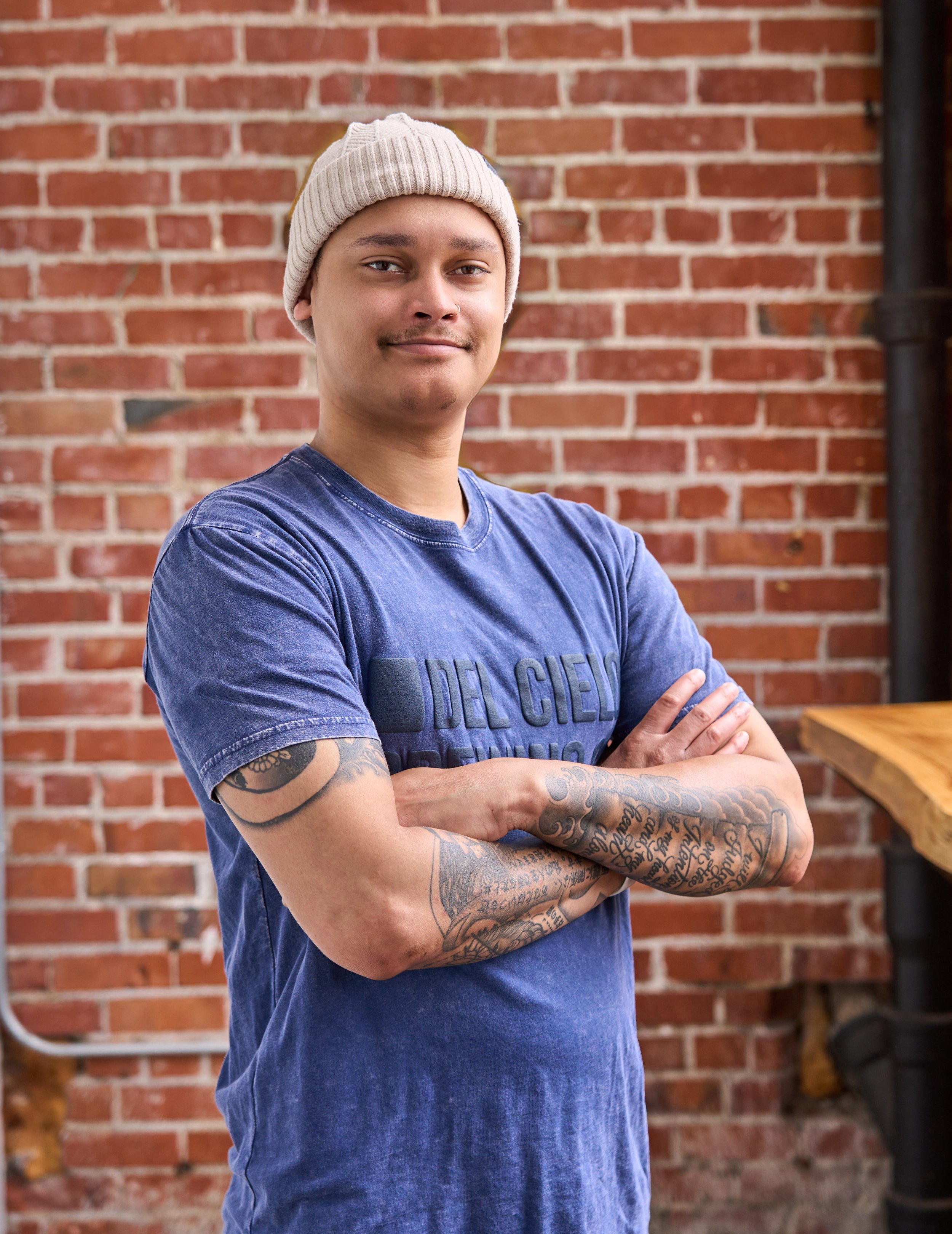 A young man with tattoos on his arms wearing a beige beanie and a blue T-shirt standing outdoors against a brick wall with a large vintage-style letter '7' sign and some greenery.