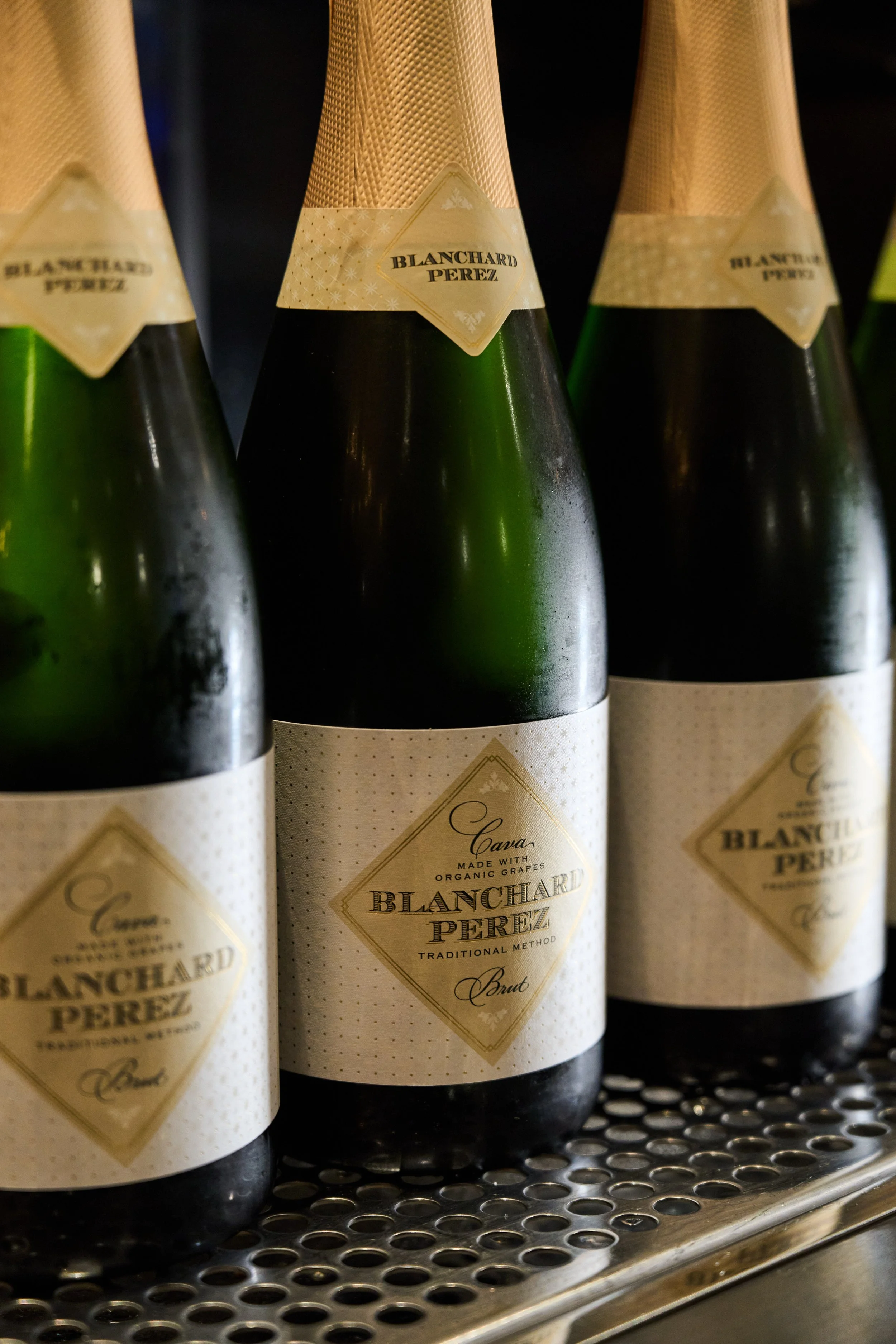 Close-up of bottles of Blancard Perez sparkling wine on a metal shelf, with beige labels and gold foil caps.