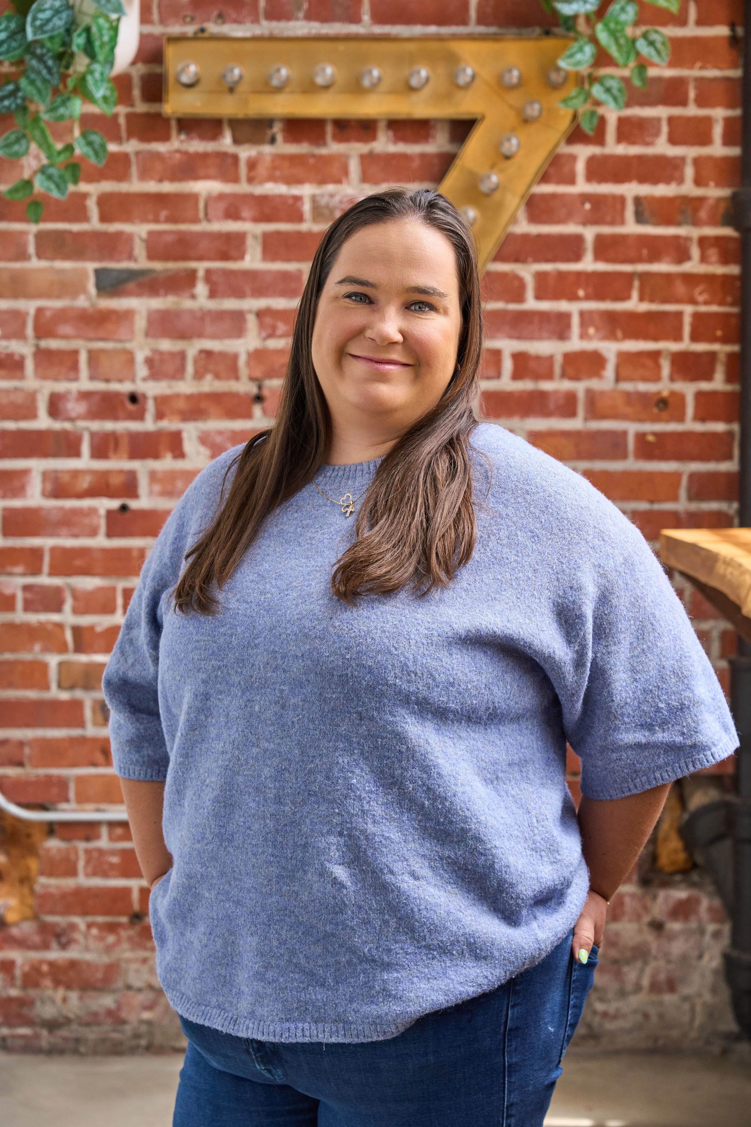 A woman with long brown hair smiling, wearing a blue sweater and jeans, standing in front of a brick wall decorated with a large lit arrow sign and green ivy.