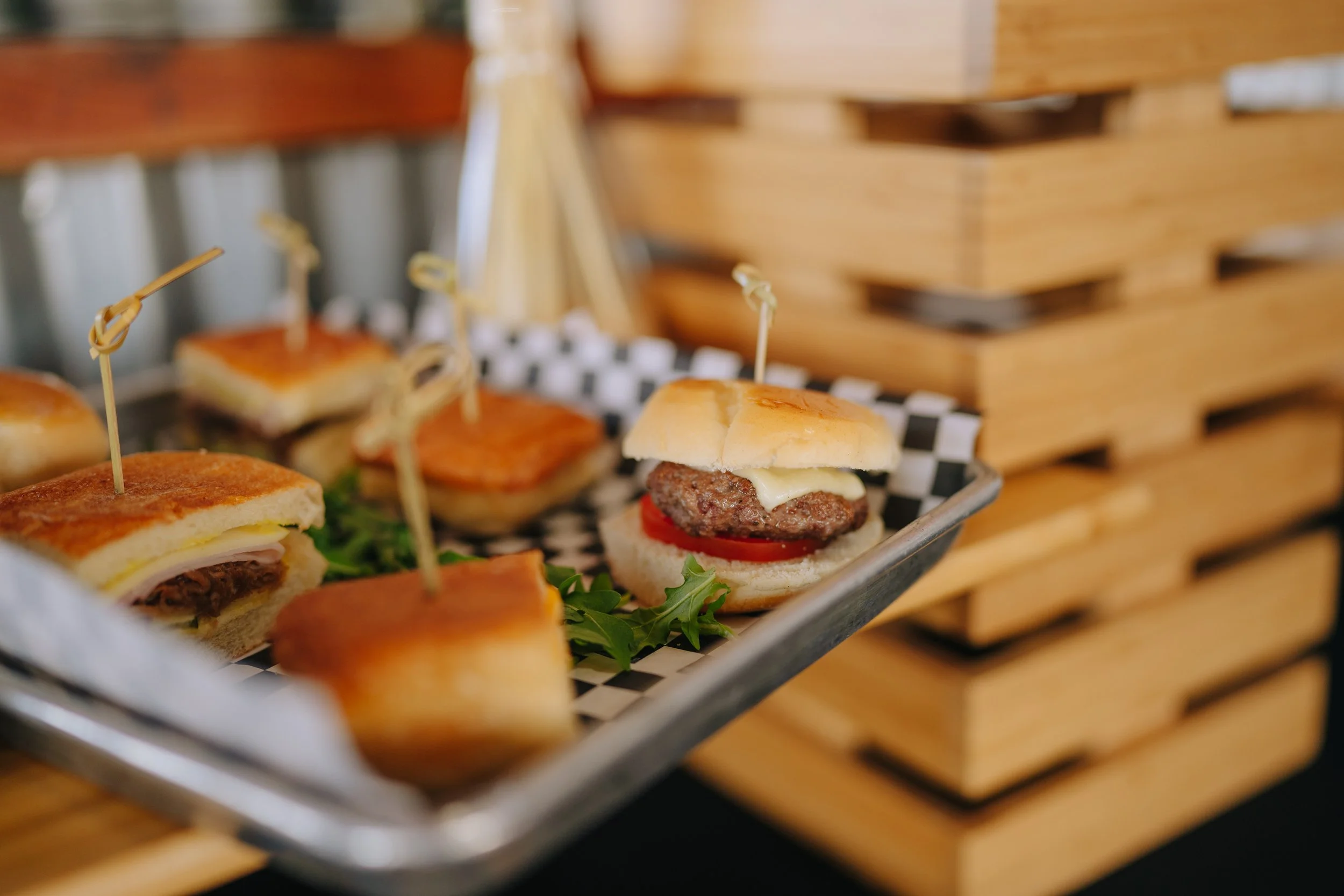 Mini sandwiches with beef patties, tomato slices, cheese, and lettuce, served on a tray lined with checkered paper.