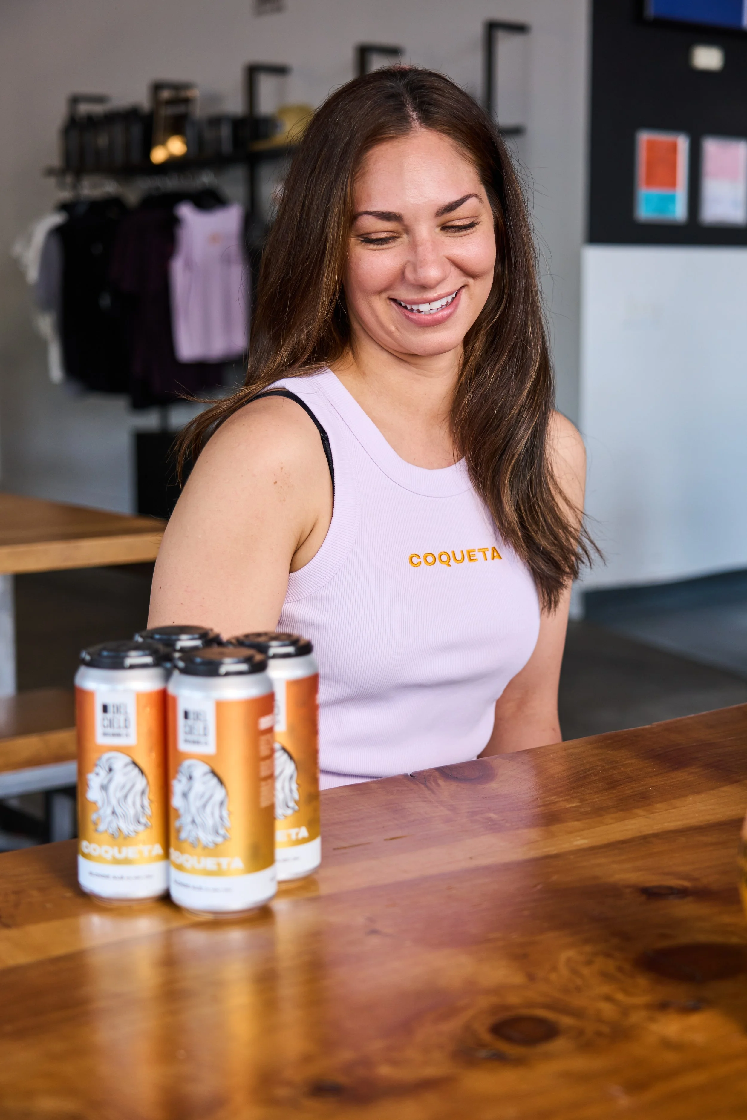 A woman with brown hair wearing a light pink tank top with 'Coqueta' written on it sitting at a wooden table with cans of Coqueta beverage in the foreground, smiling with eyes closed in an indoor setting.