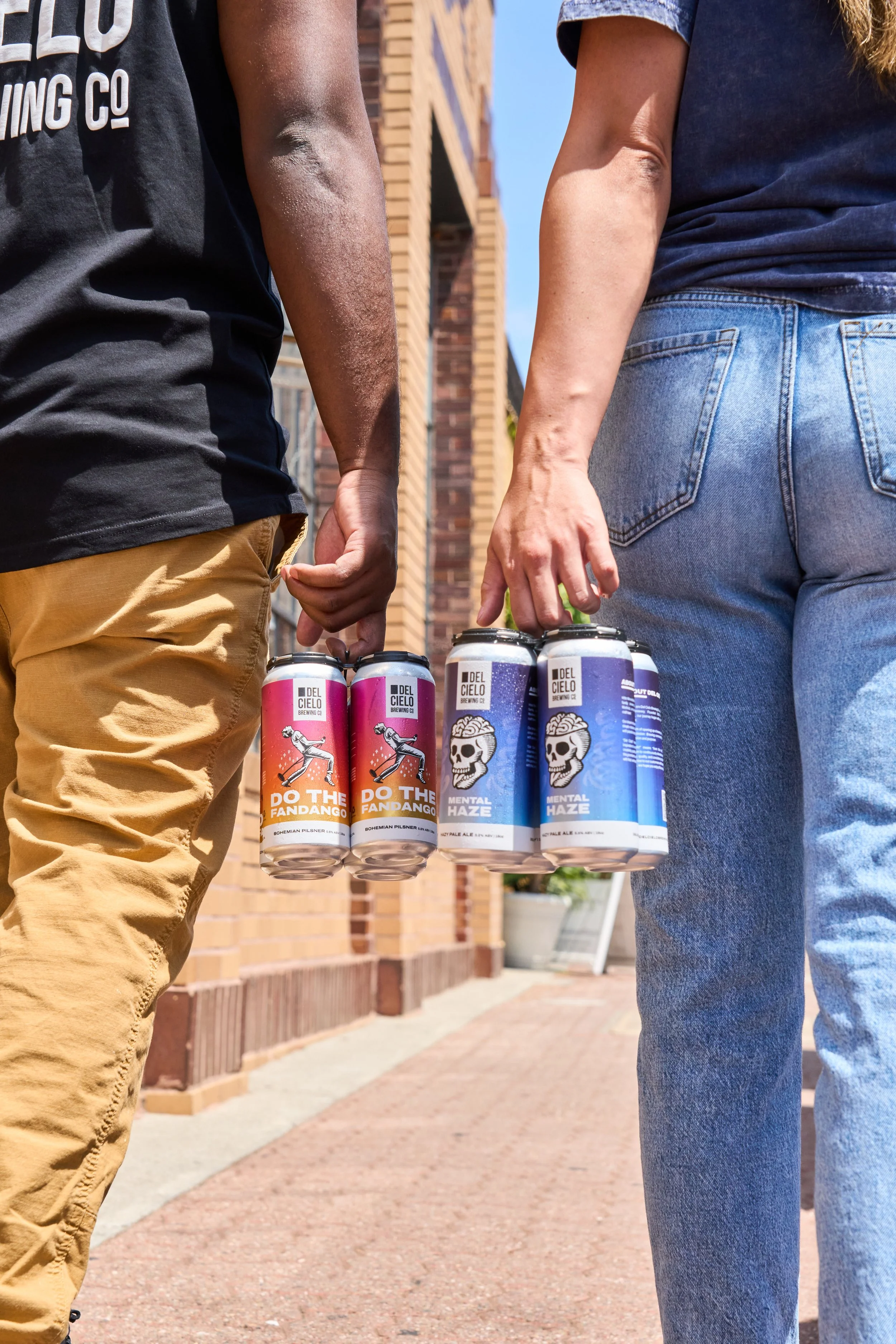Two people holding cans of beer during daytime, standing outdoors in front of a brick building.