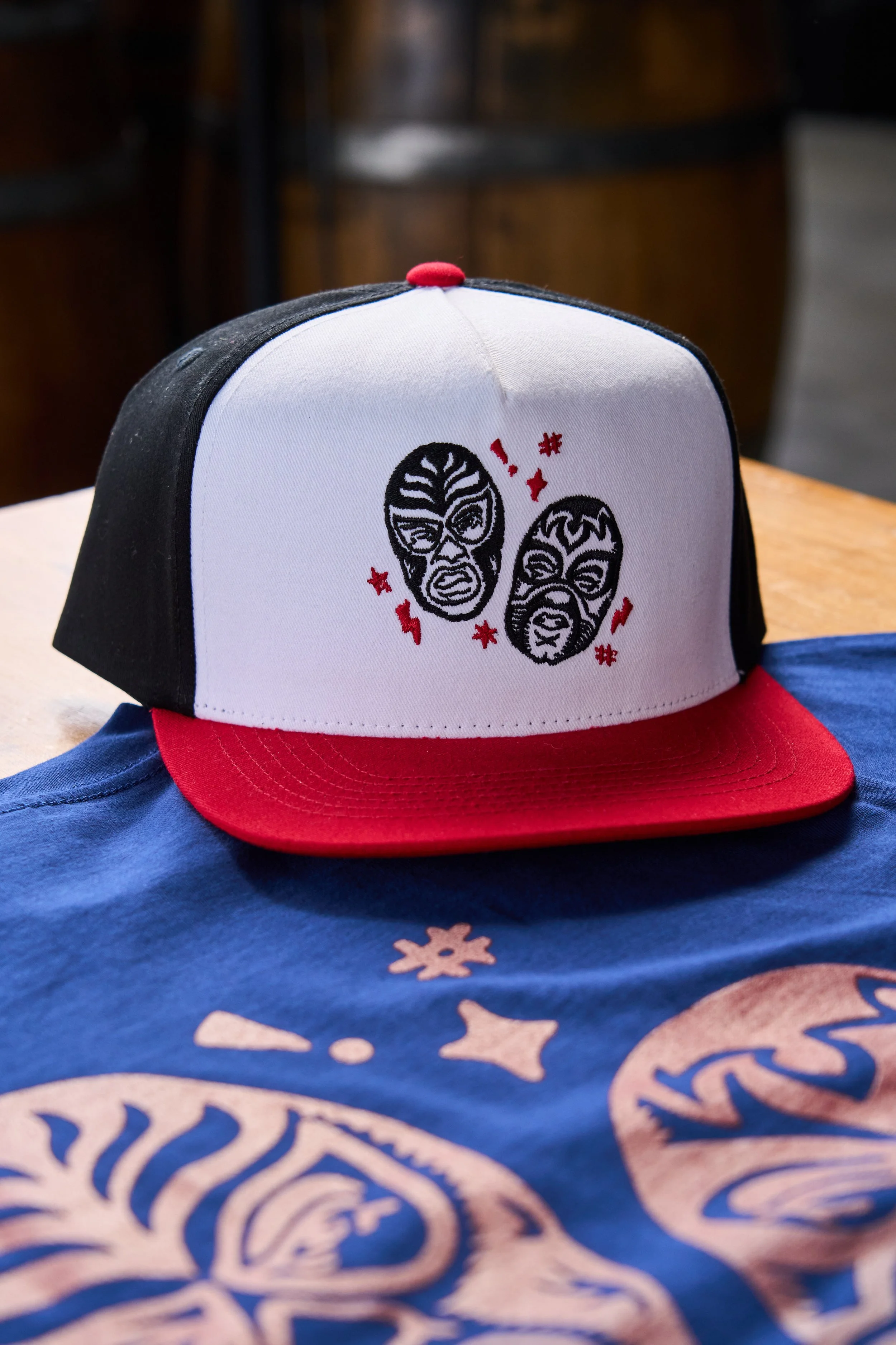 A white trucker hat with a red brim and black mesh back, decorated with embroidered lucha libre masks and red accents on the front.