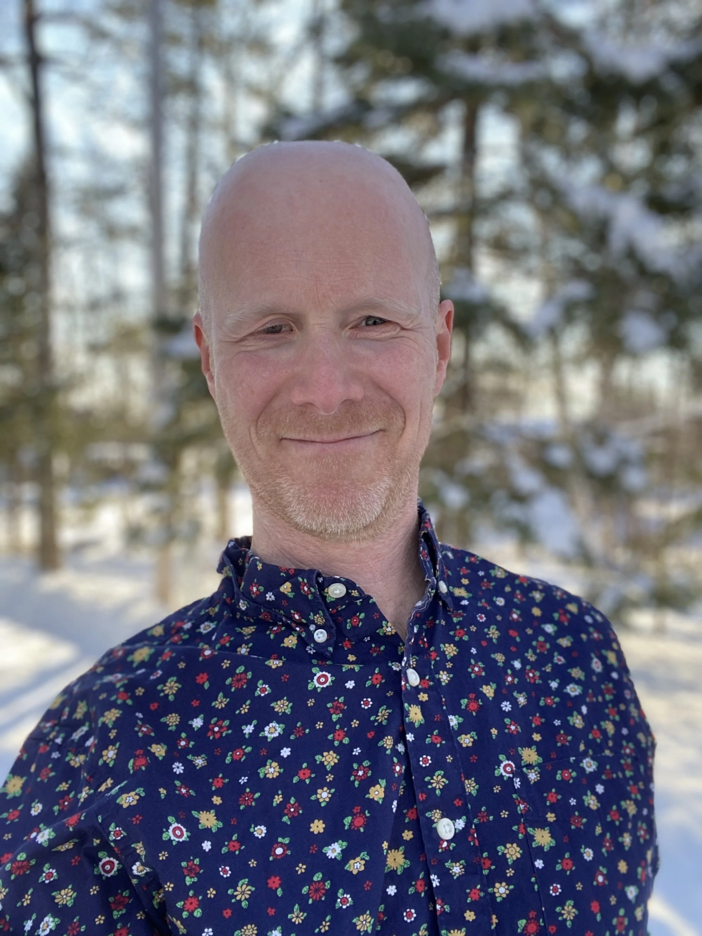 A man with a bald head and light facial hair smiling outdoors in a snowy wooded area, wearing a dark blue shirt with a colorful floral pattern.