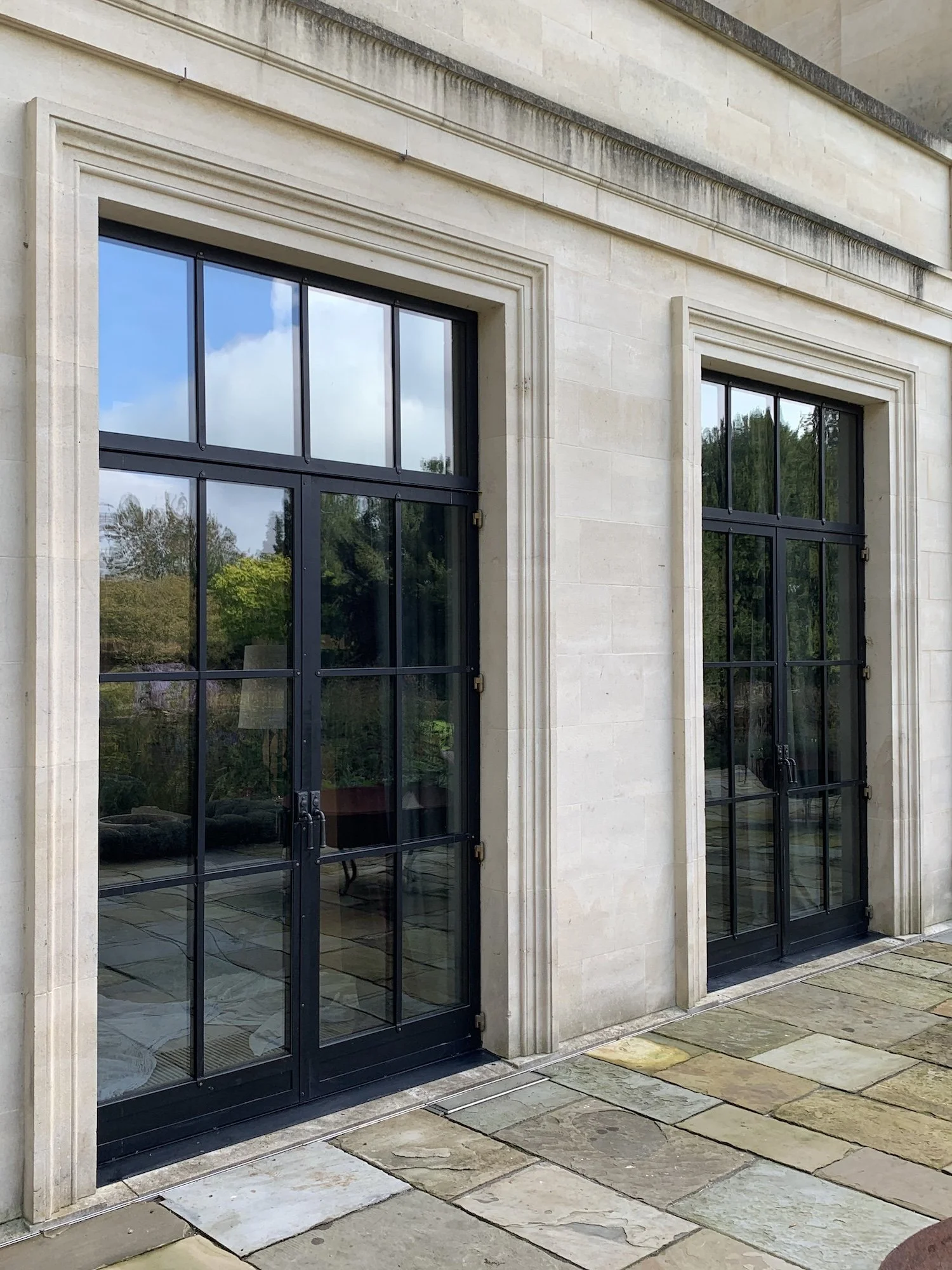 Exterior of a building with two large black-framed glass doors and a light-colored stone facade, showing a view of a garden with trees reflected in the glass.