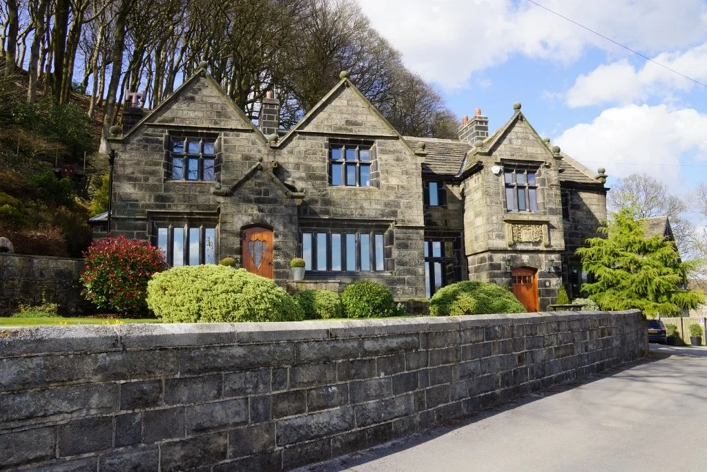 Stone-built mansion with multiple gabled roofs, large windows, wooden doors, and surrounding shrubbery, set against a backdrop of trees and a blue sky.