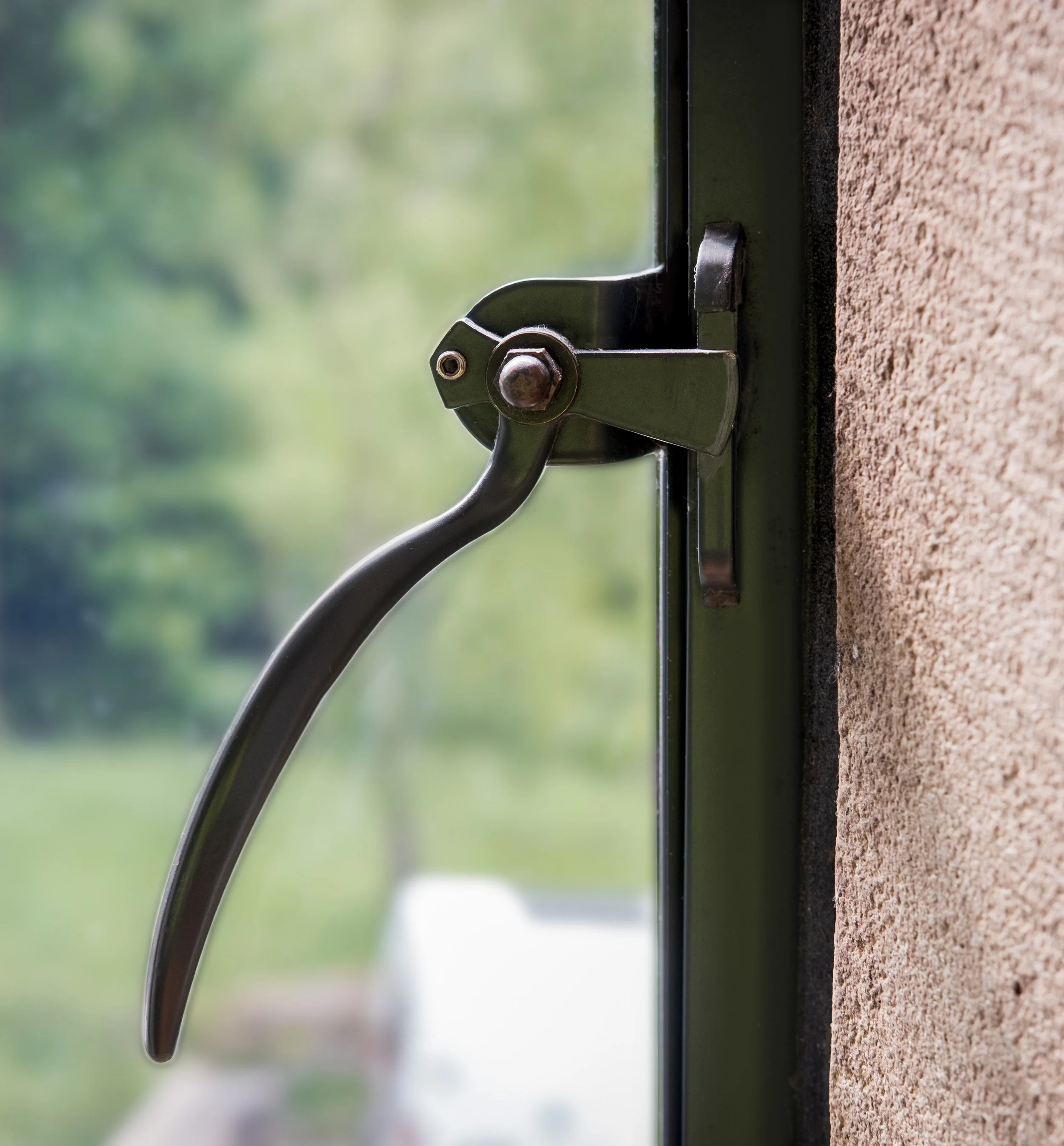 Close-up of a black window latch on a glass window with a blurred green outdoor background and a textured brown window sill.