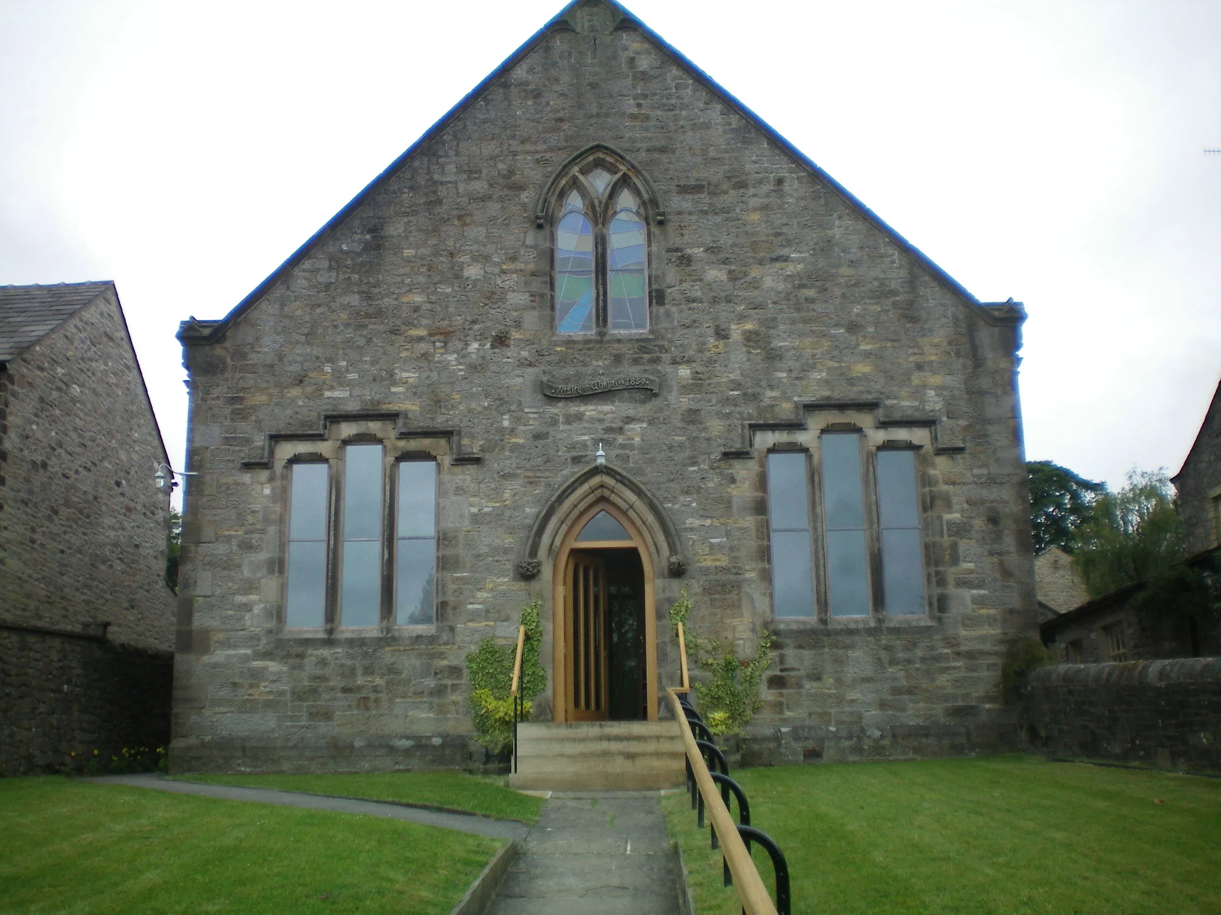 Front view of a stone church with a stained glass window on the gable end and wooden door, surrounded by a grassy lawn and walkways.