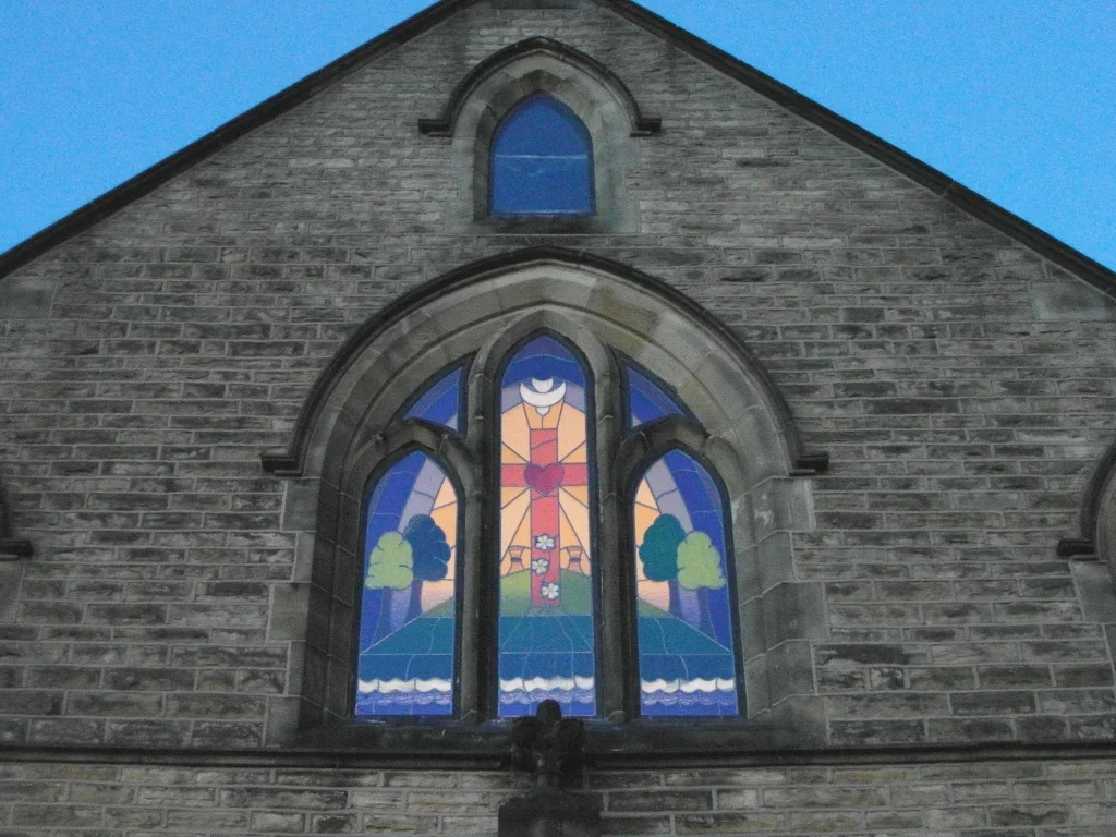 Colorful stained glass church window with a heart and cross design, surrounded by trees and sky.