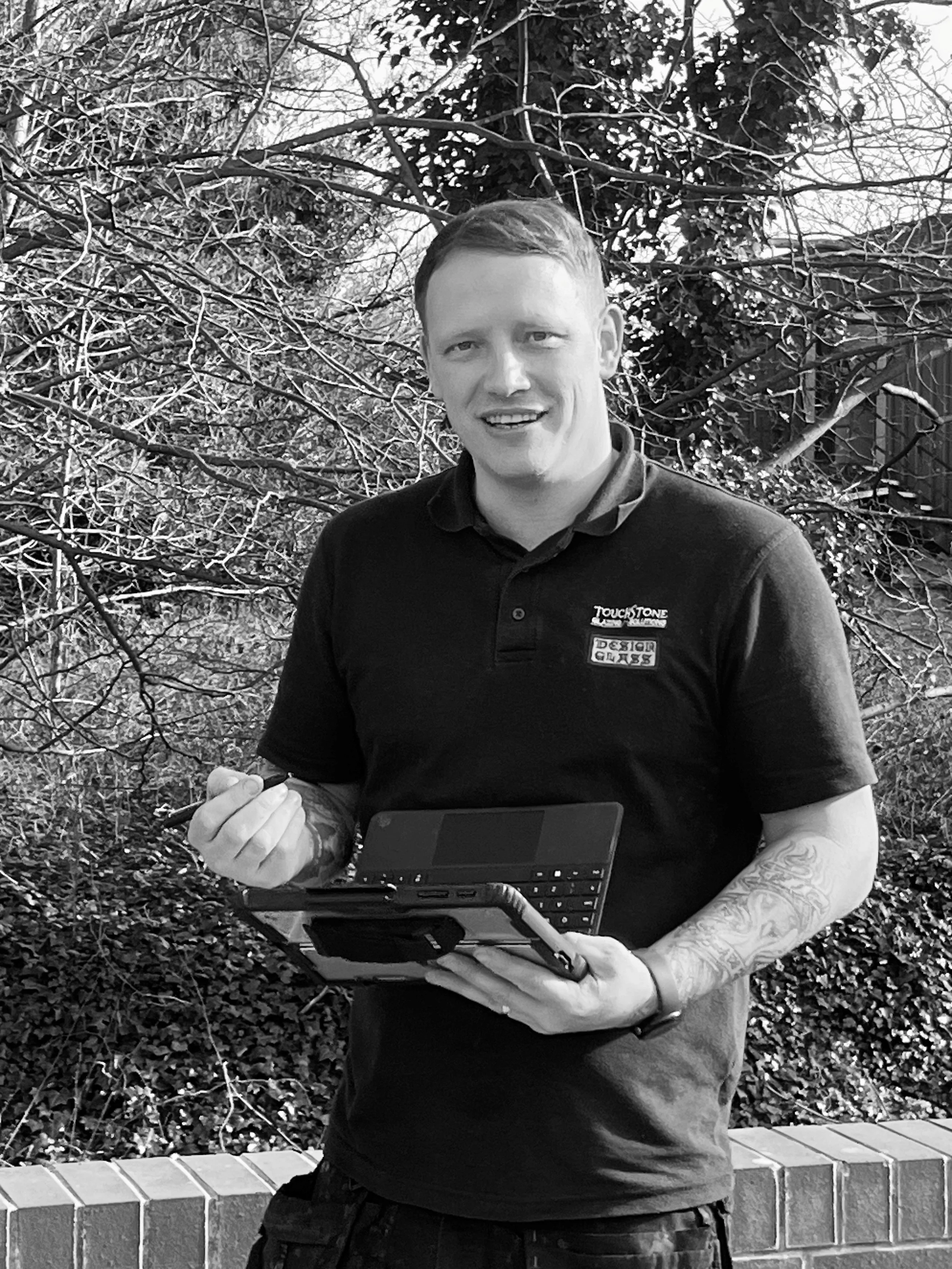 A man standing outdoors, holding a laptop and stylus, smiling. He has tattoos on his right arm, short hair, and is wearing a dark polo shirt with logos. Background includes trees, bushes, and a brick wall.