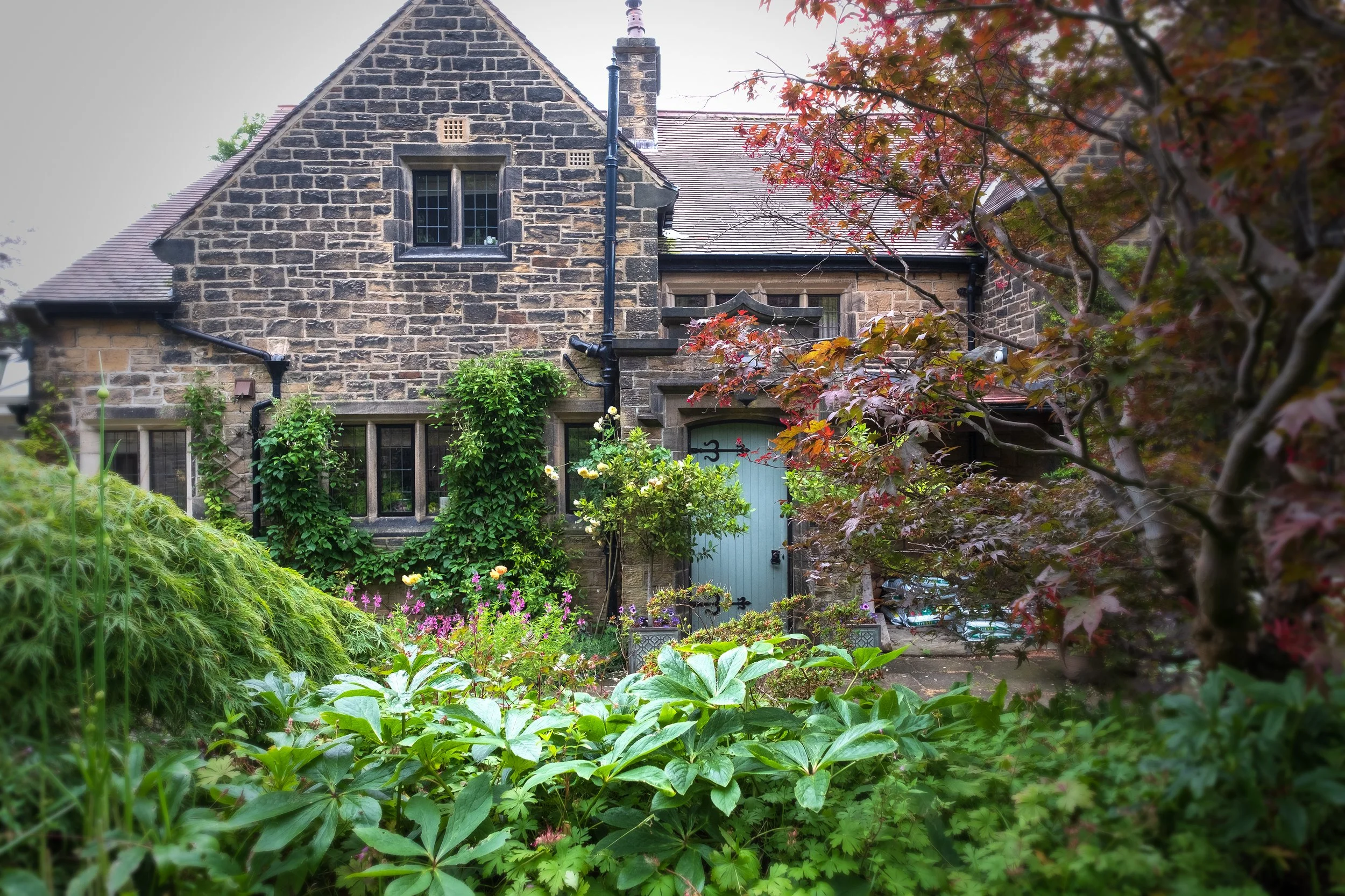 A stone house with a garden in front, including various plants and trees with red and green leaves.