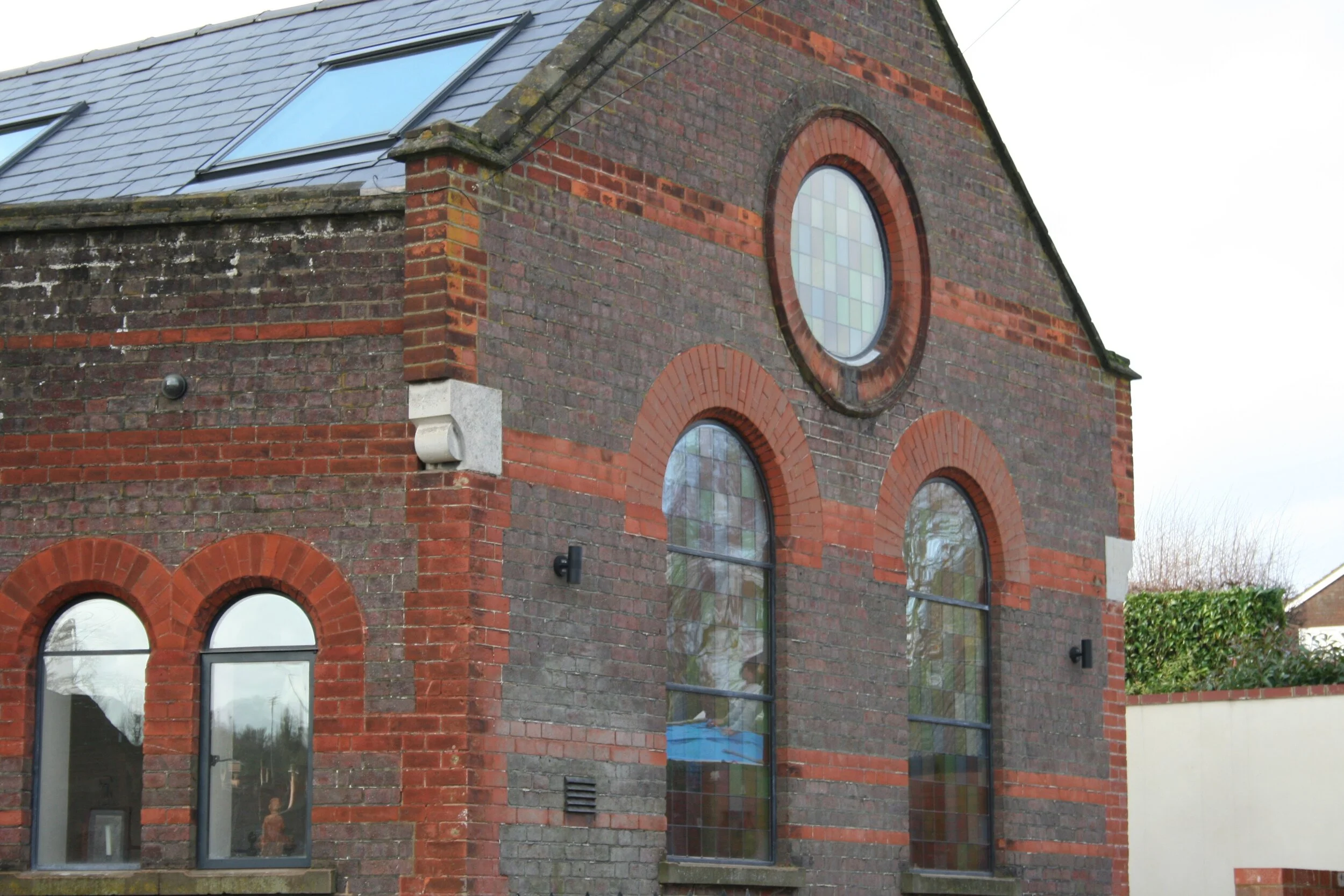 The image shows the exterior of a brick building with large arched and rectangular windows, some of which have stained glass. The roof is sloped with tiles and features skylights.