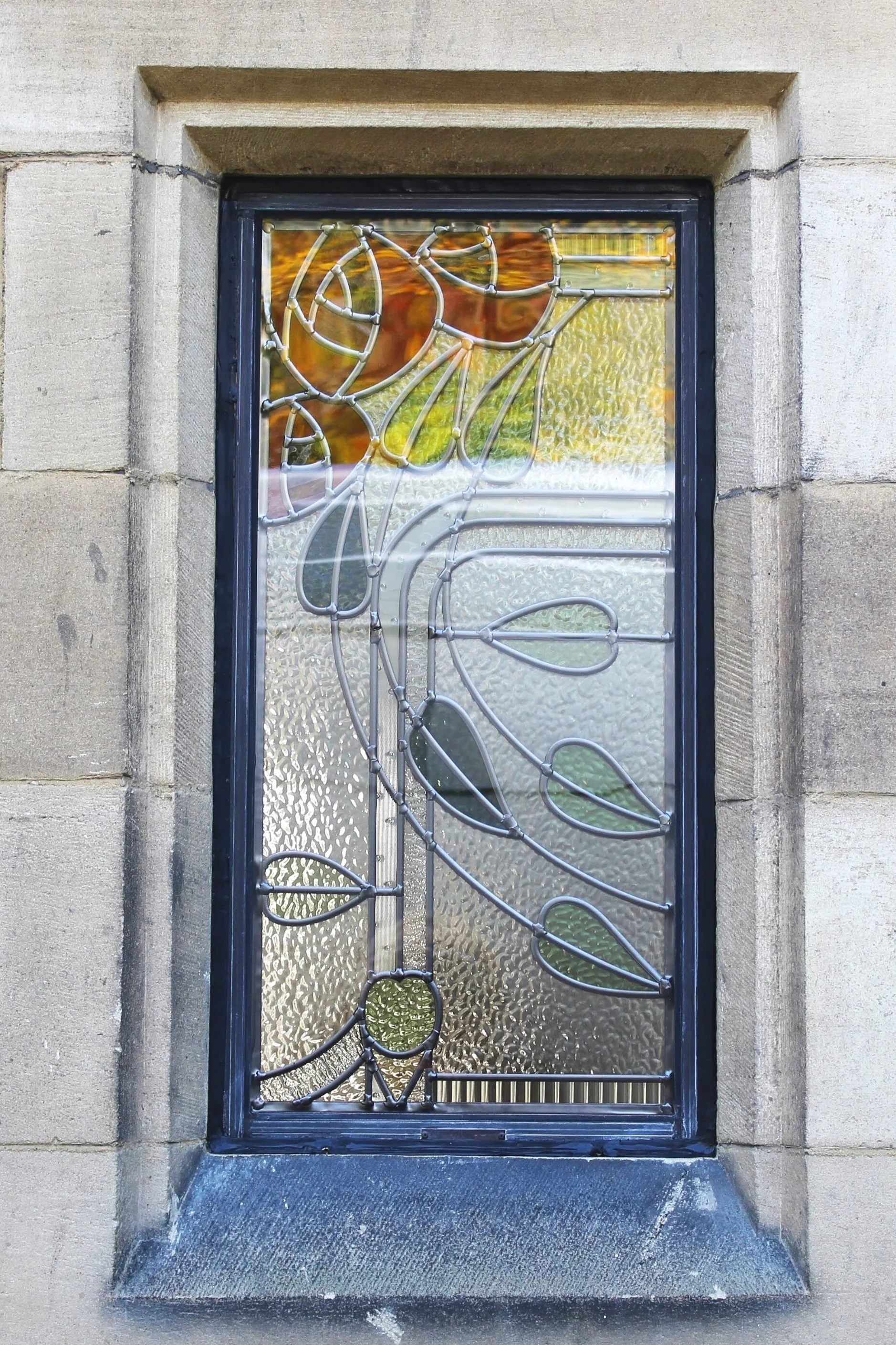 Decorative stained glass window featuring a floral design with leaves and a flower, set in a stone wall.