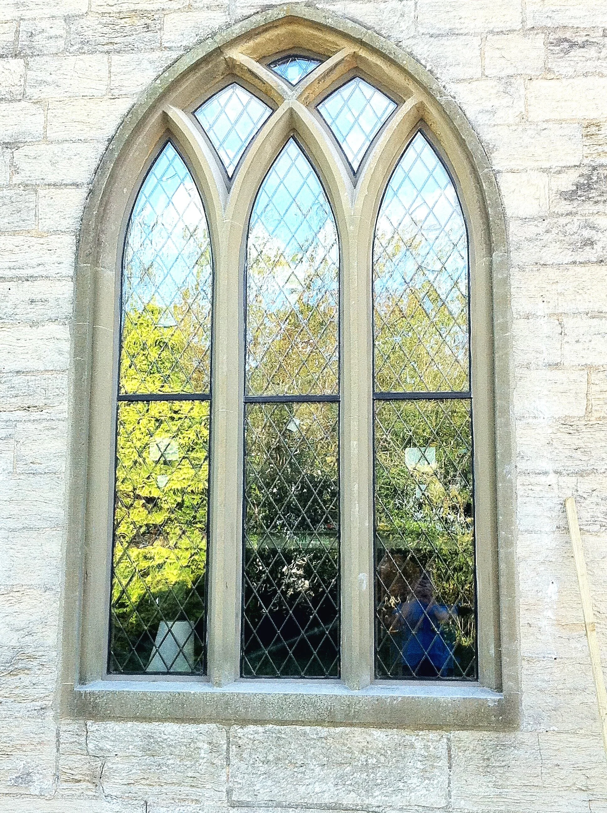 A tall, arched, Gothic-style window with diamond-shaped glass panes set in a stone frame, on a stone wall of a building.
