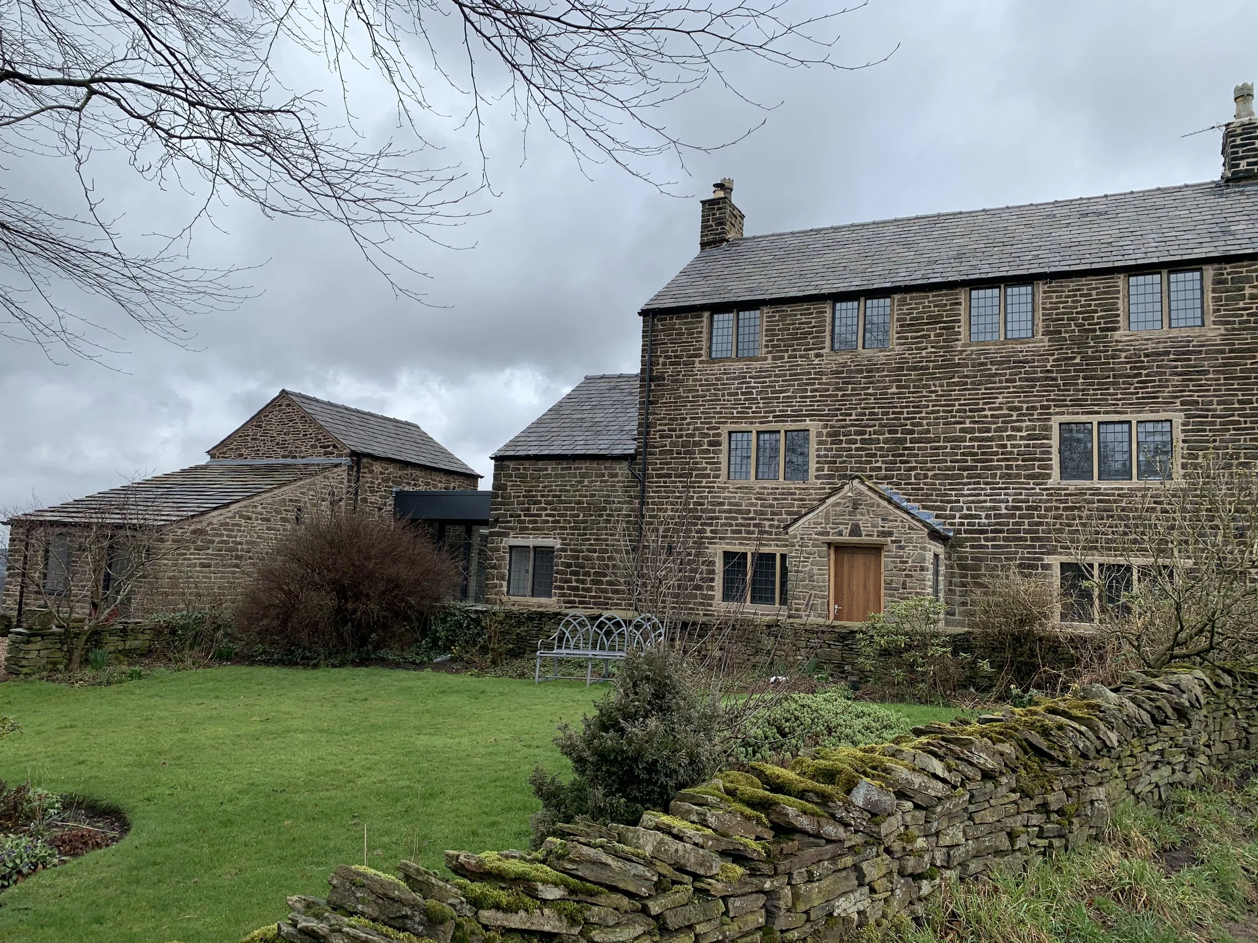 A traditional stone cottage with multiple windows, a small porch, and a slate roof, surrounded by a lush green garden and a stone wall, under a cloudy sky.