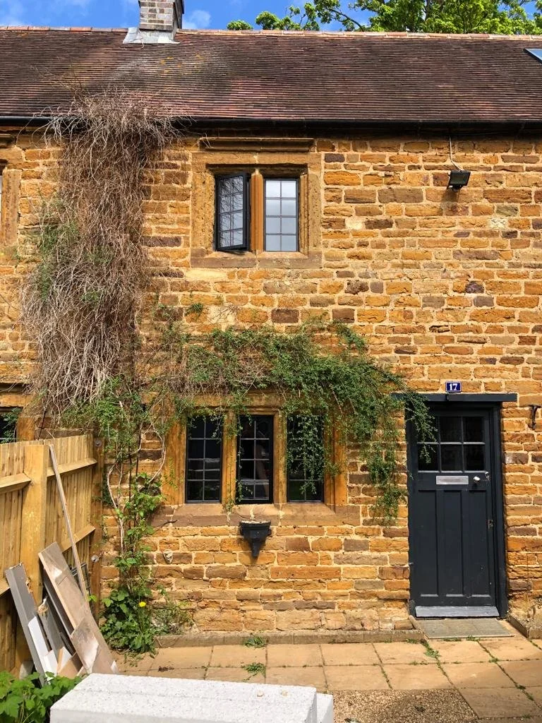 Brick house with a black door and windows, some greenery over the window, a small window above, and a vine growing on the left side.