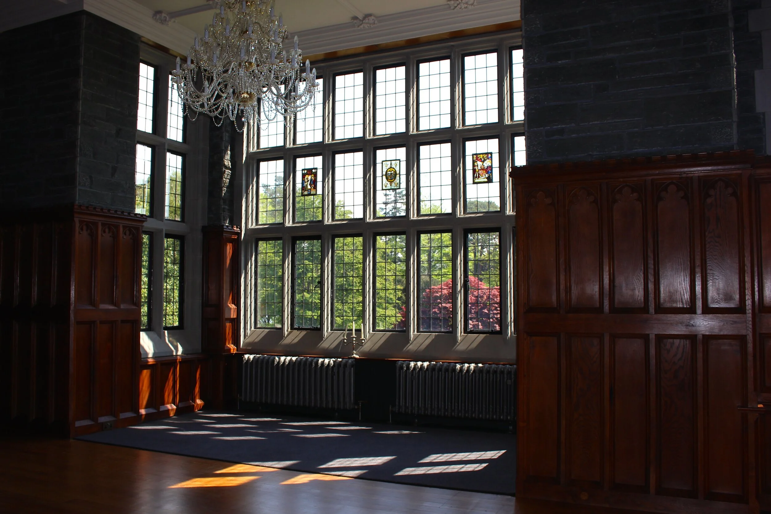 Interior of a room with large stained glass windows, wooden paneling, a crystal chandelier, and sunlight casting shadows on the floor.