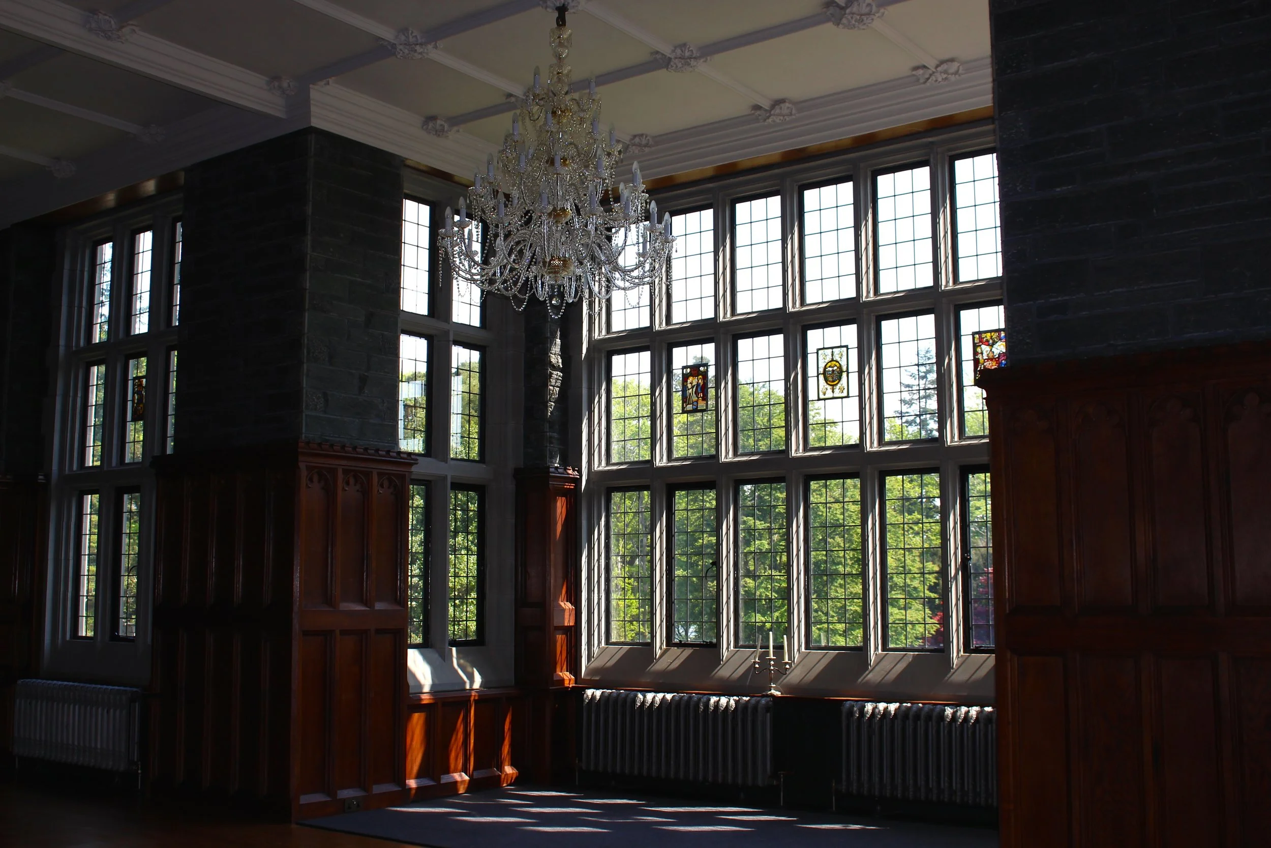 Interior of a historic building with large stained glass windows, a crystal chandelier, wooden paneling, and a black radiator beneath the windows, with sunlight streaming in.