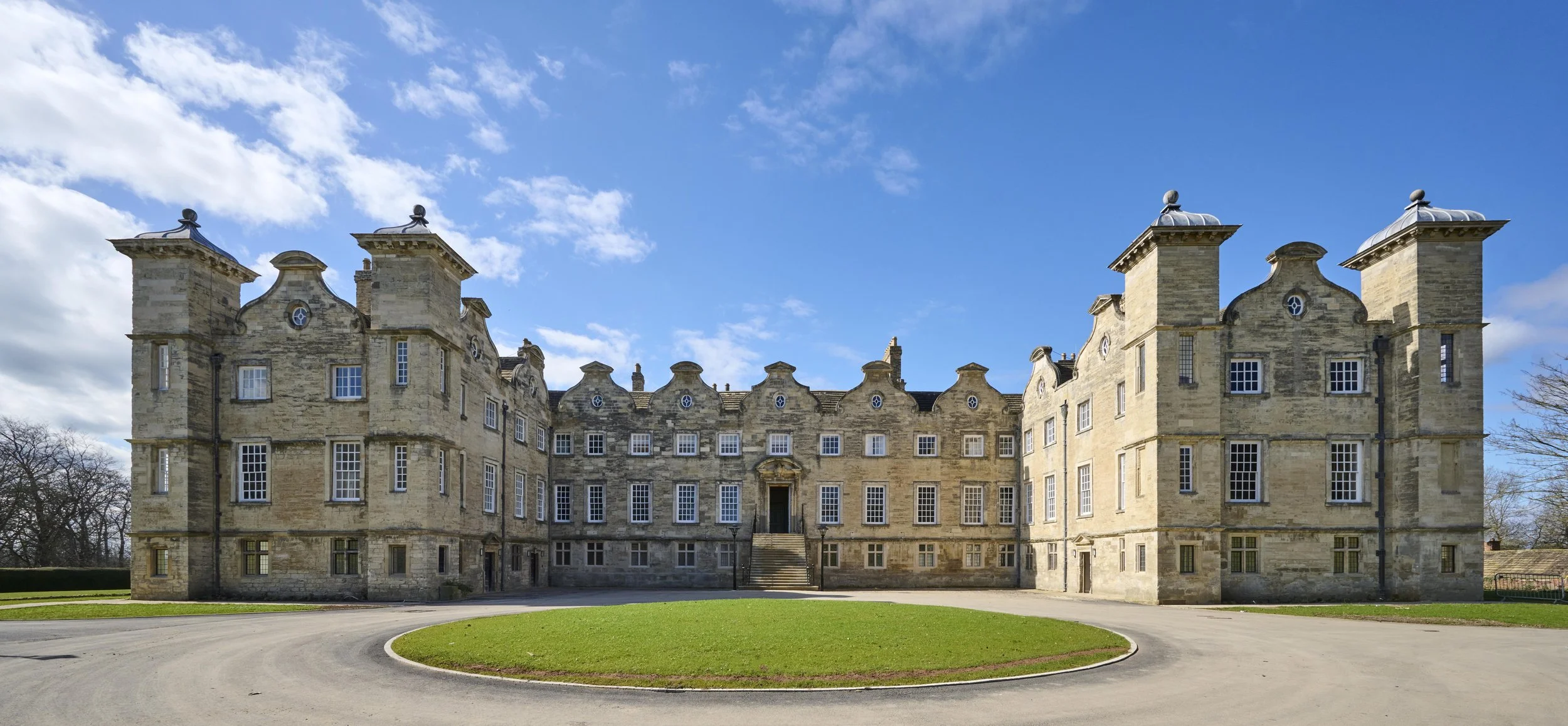 A large historic stone castle with multiple turrets and numerous windows, set against a blue sky with scattered clouds, and surrounded by a circular driveway with a grassy area in front.