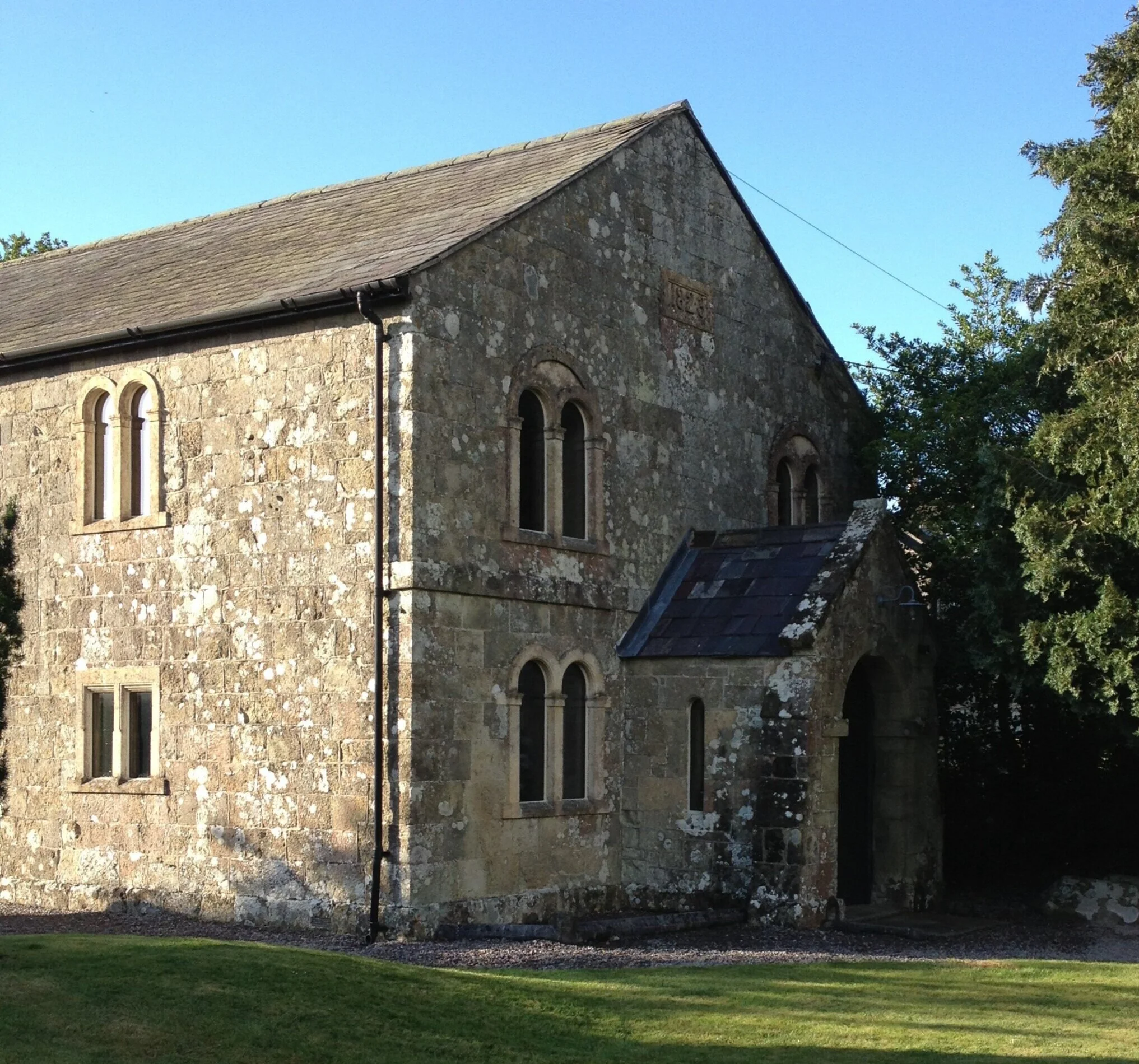 An old stone church with arched windows and a small entrance, surrounded by trees and grass on a sunny day.