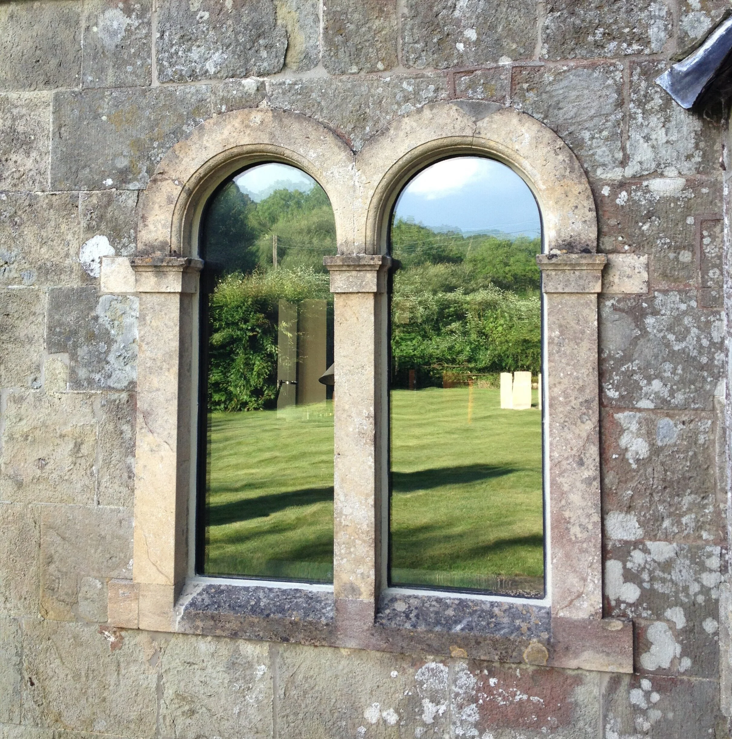 Two arched stone-framed windows on a stone wall reflecting a green lawn and trees, with a cloudy sky visible in the background.