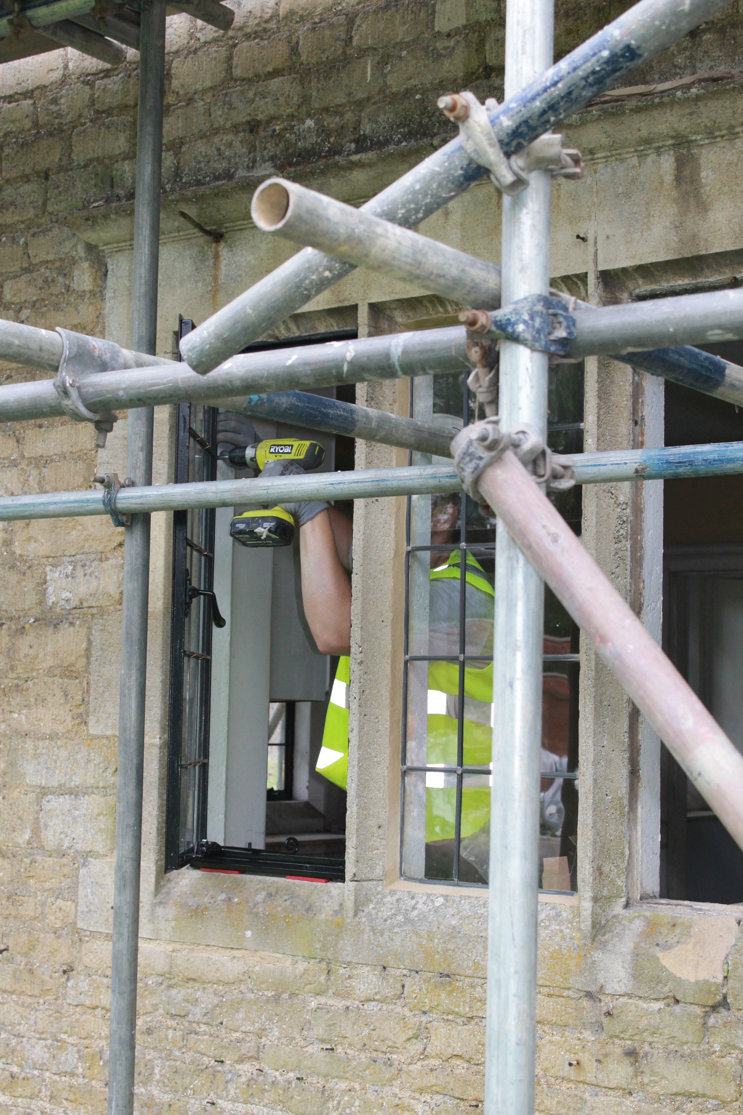 A construction worker wearing a yellow safety vest and gloves is using a cordless drill on a window frame surrounded by scaffolding on a brick building.