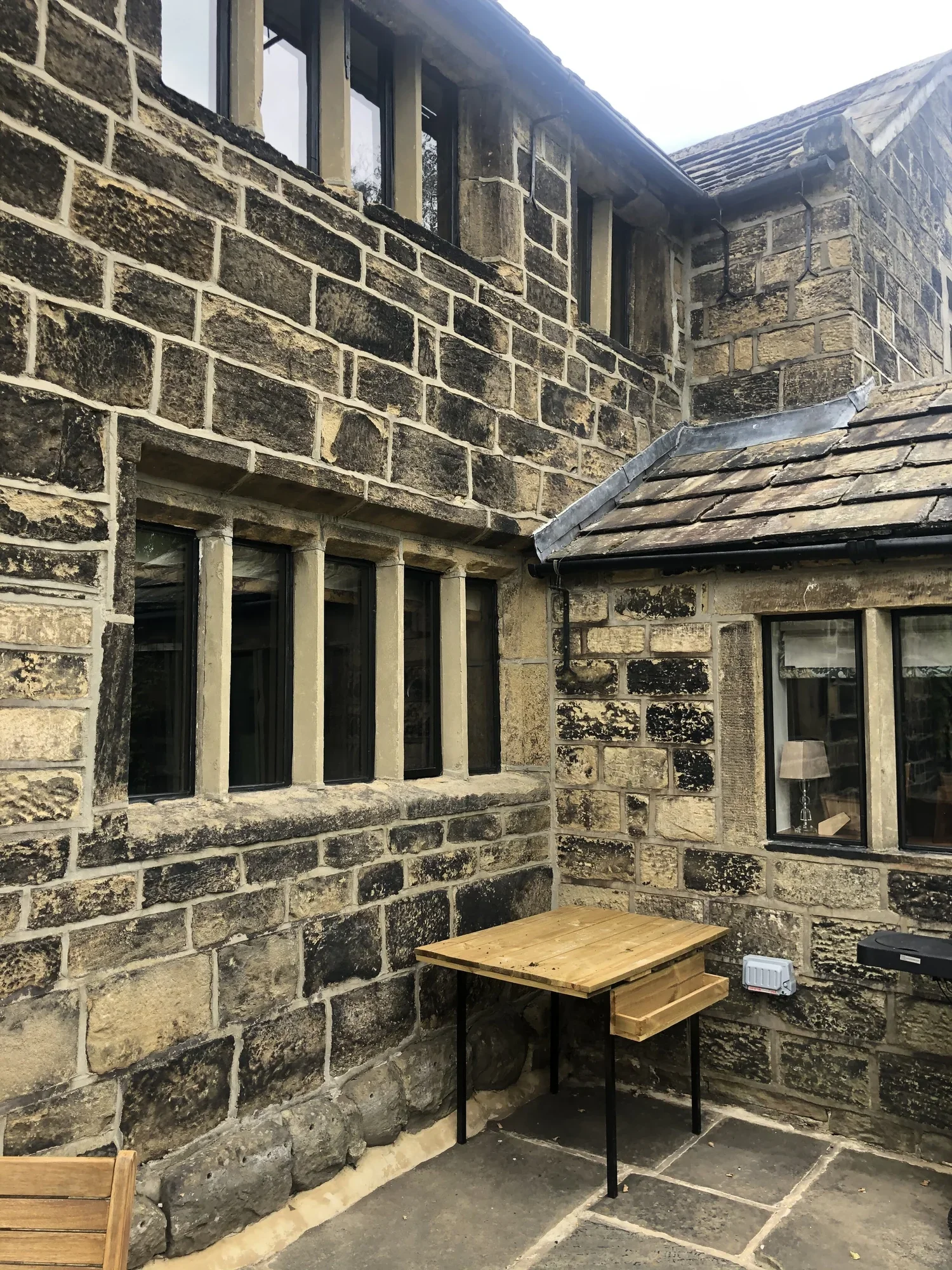 A stone building with black-framed windows and a small outdoor wooden table. There is a lamp and a gray object on the ground. The building has a sloped roof and a rain gutter.
