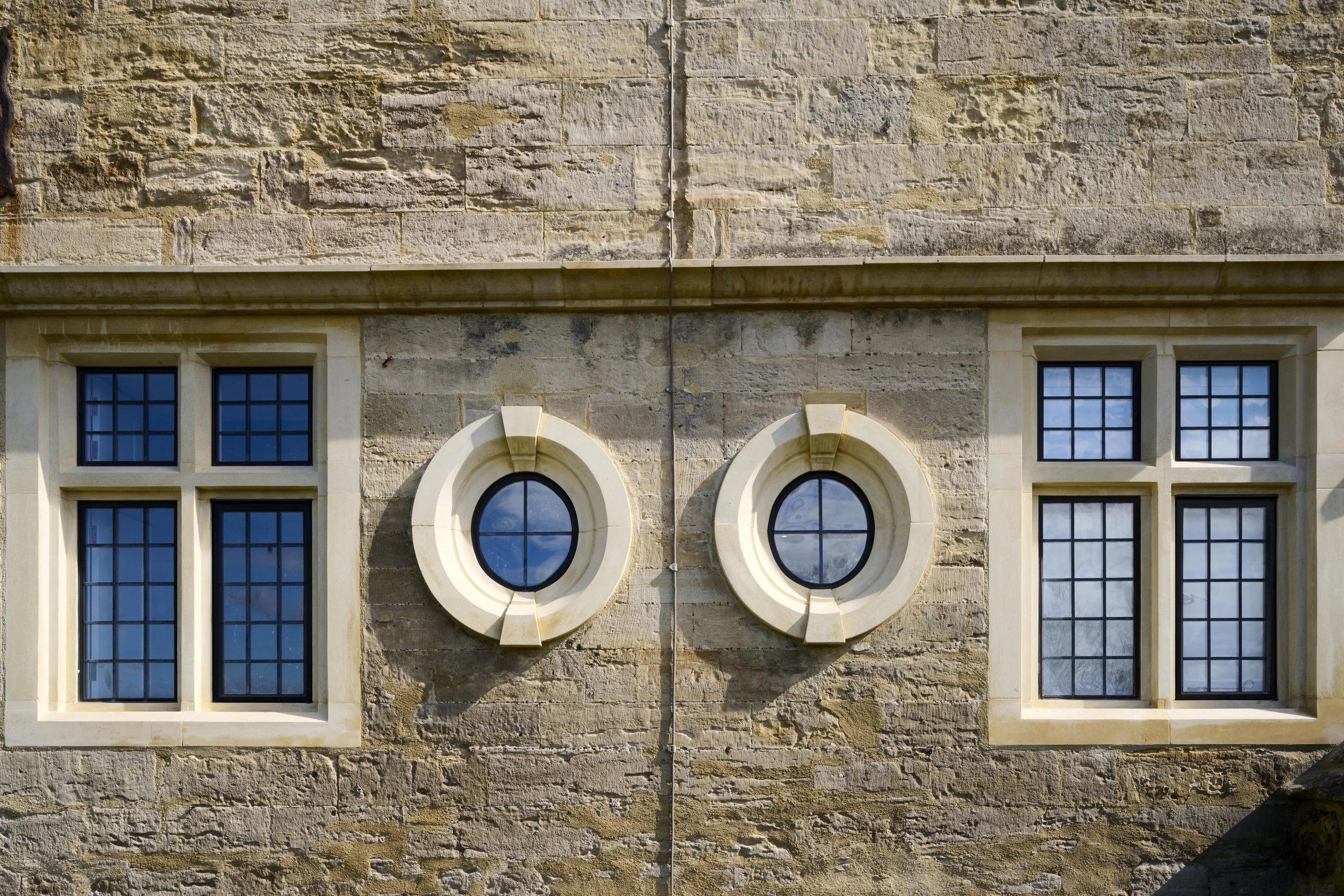 Stone building facade with four black window panes and two circular decorative elements. Orange pipe runs vertically down the center of the wall.