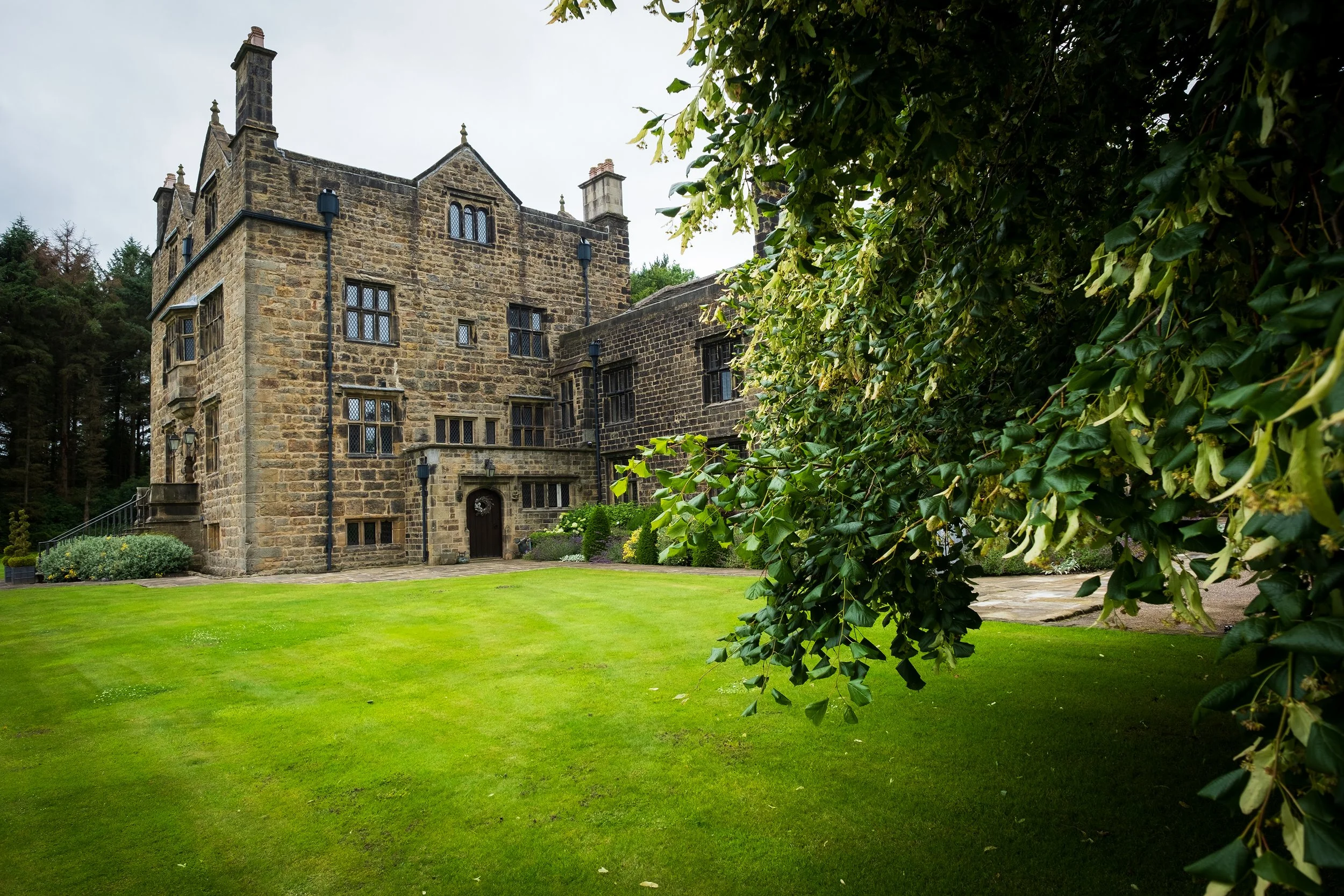A large stone castle with multiple windows and a dark door surrounded by a well-maintained green lawn and trees, partially hidden by green foliage on the right.