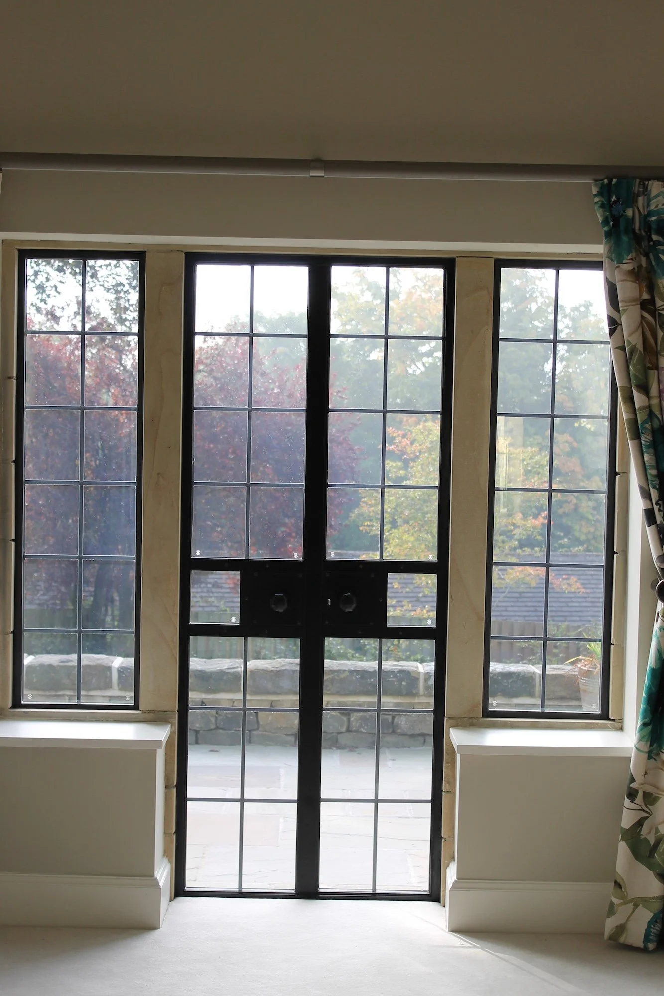 Large window with multiple panes and a black door leading outside to a garden with trees and stone borders, surrounded by cream-colored walls and baseboards, with floral curtains on the right.