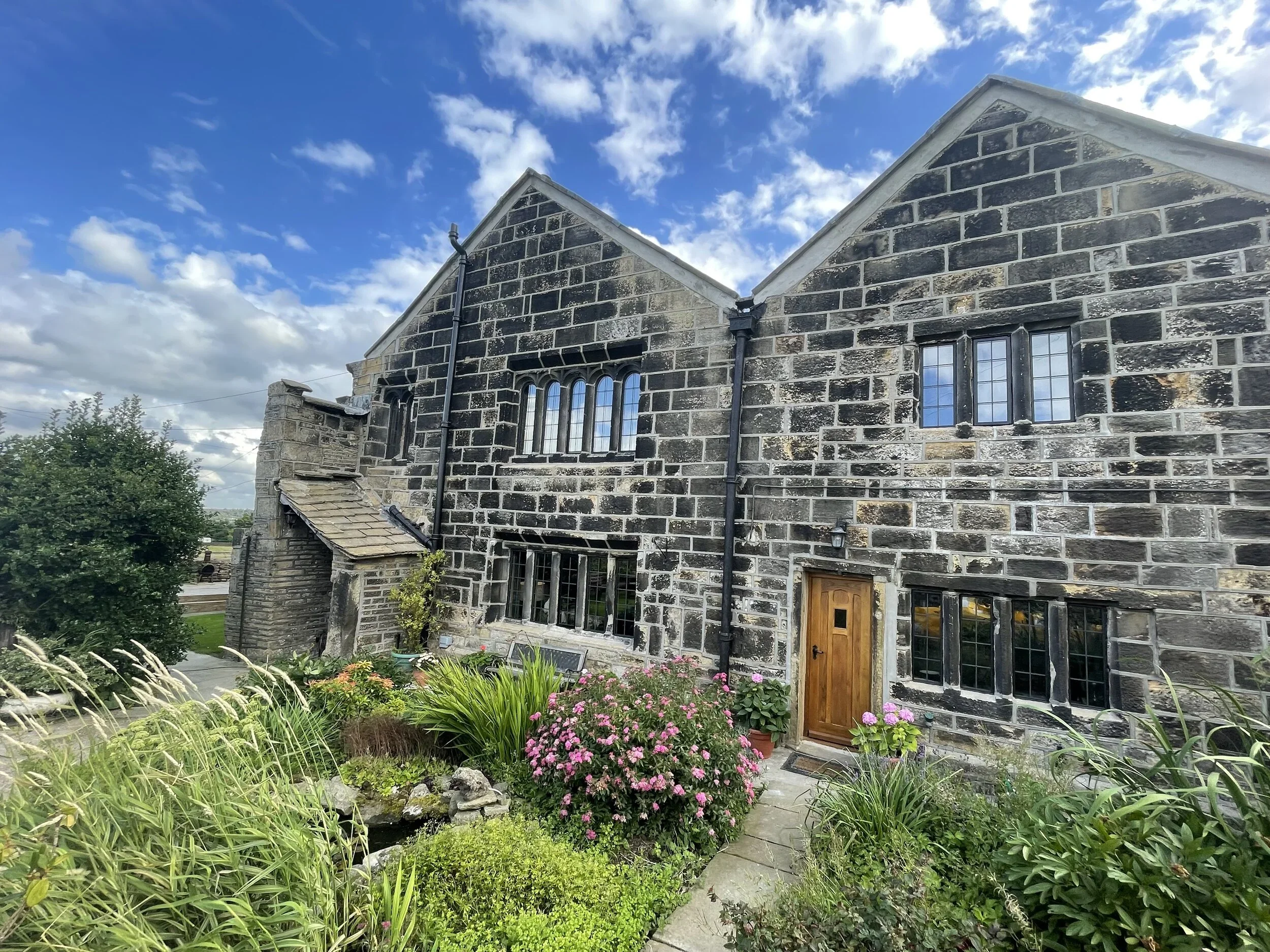 A stone house with a steep roof, black gutters, and large windows, surrounded by a garden with pink flowers and greenery, under a partly cloudy sky.