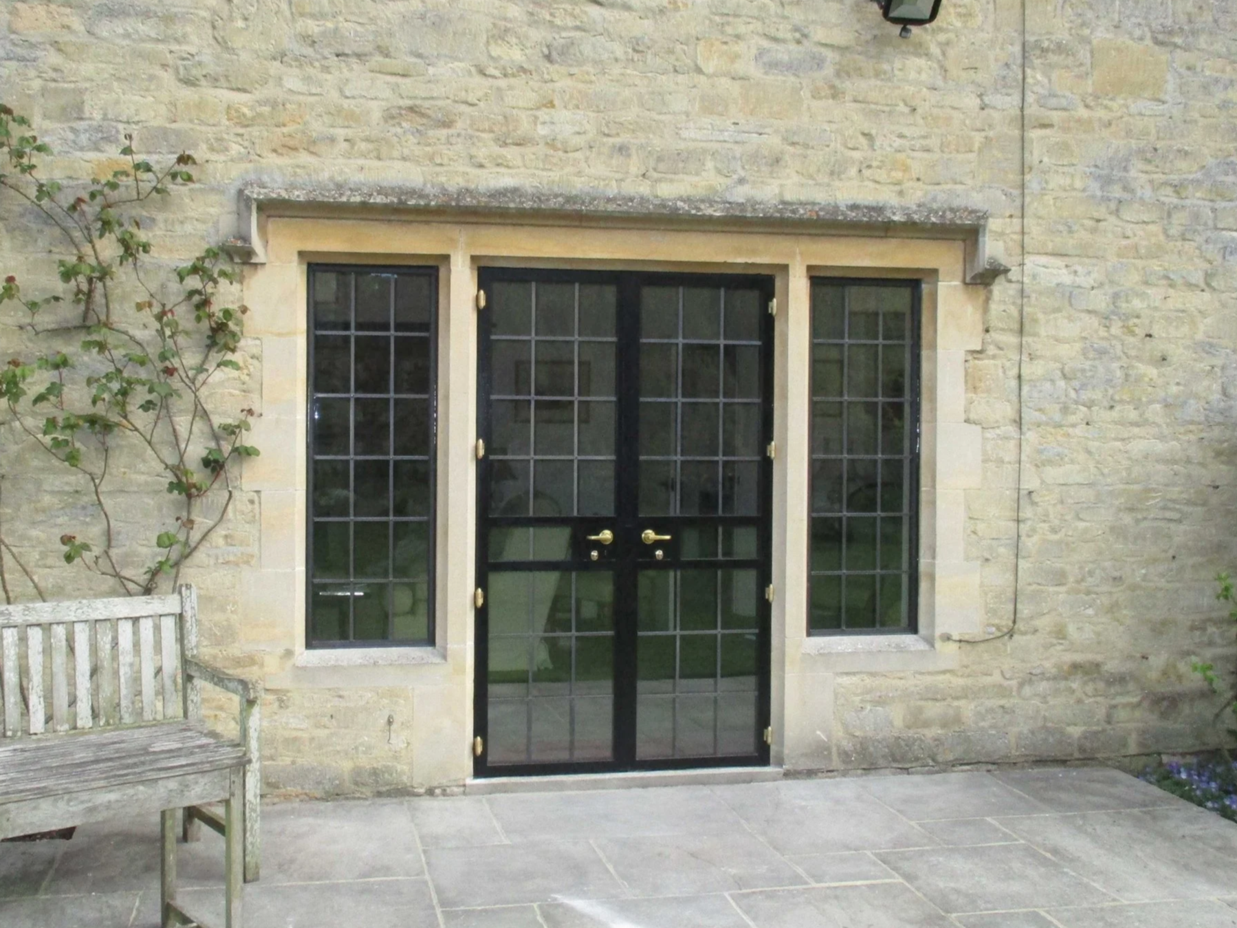 A black glass door with two handles and two side windows, set in a stone wall, with a wooden bench to the left and plants along the wall.