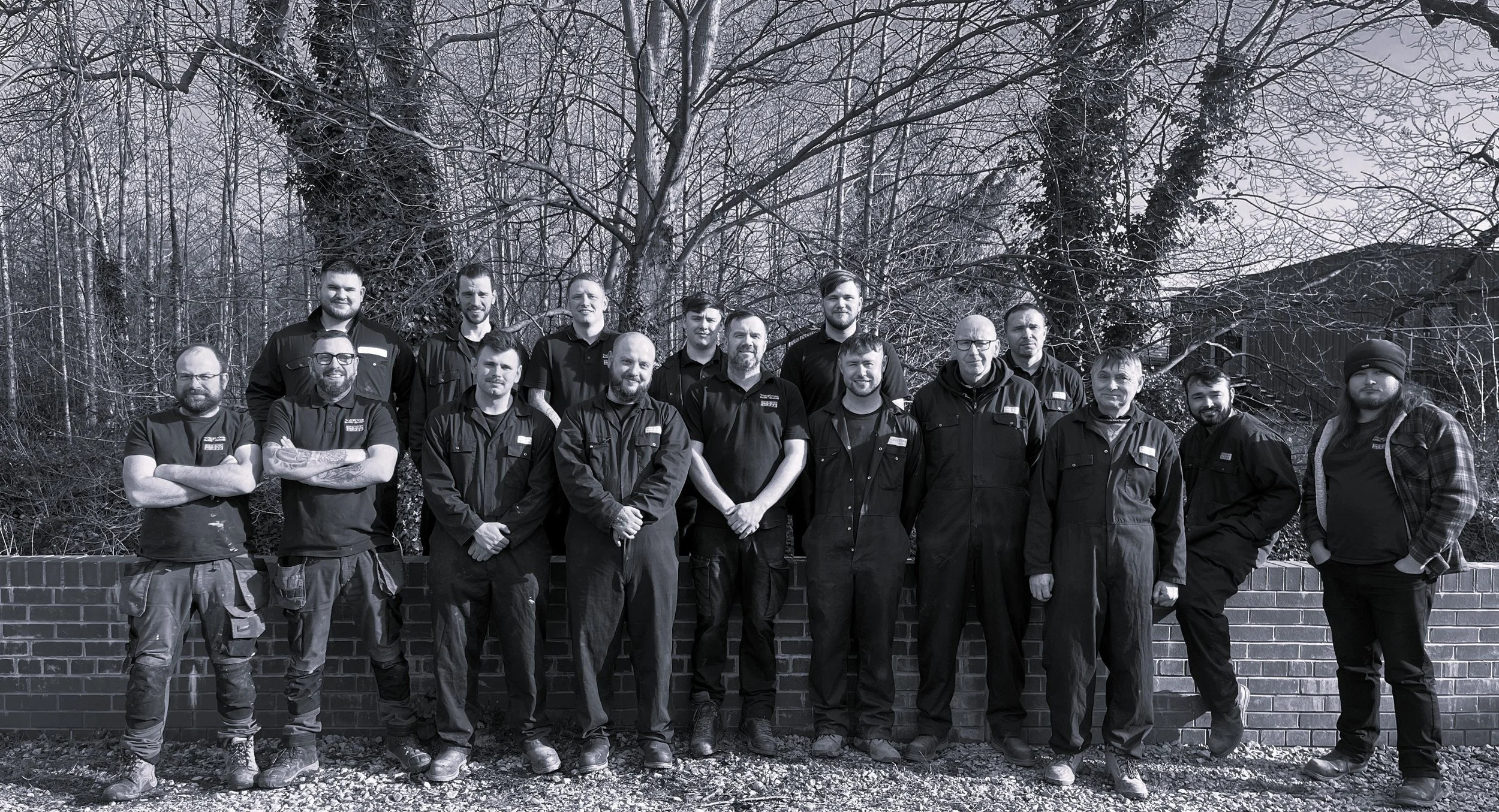 Group of fifteen men standing outdoors against a brick wall and leafless trees, dressed in work uniforms, some with arms crossed, with a few smiling.