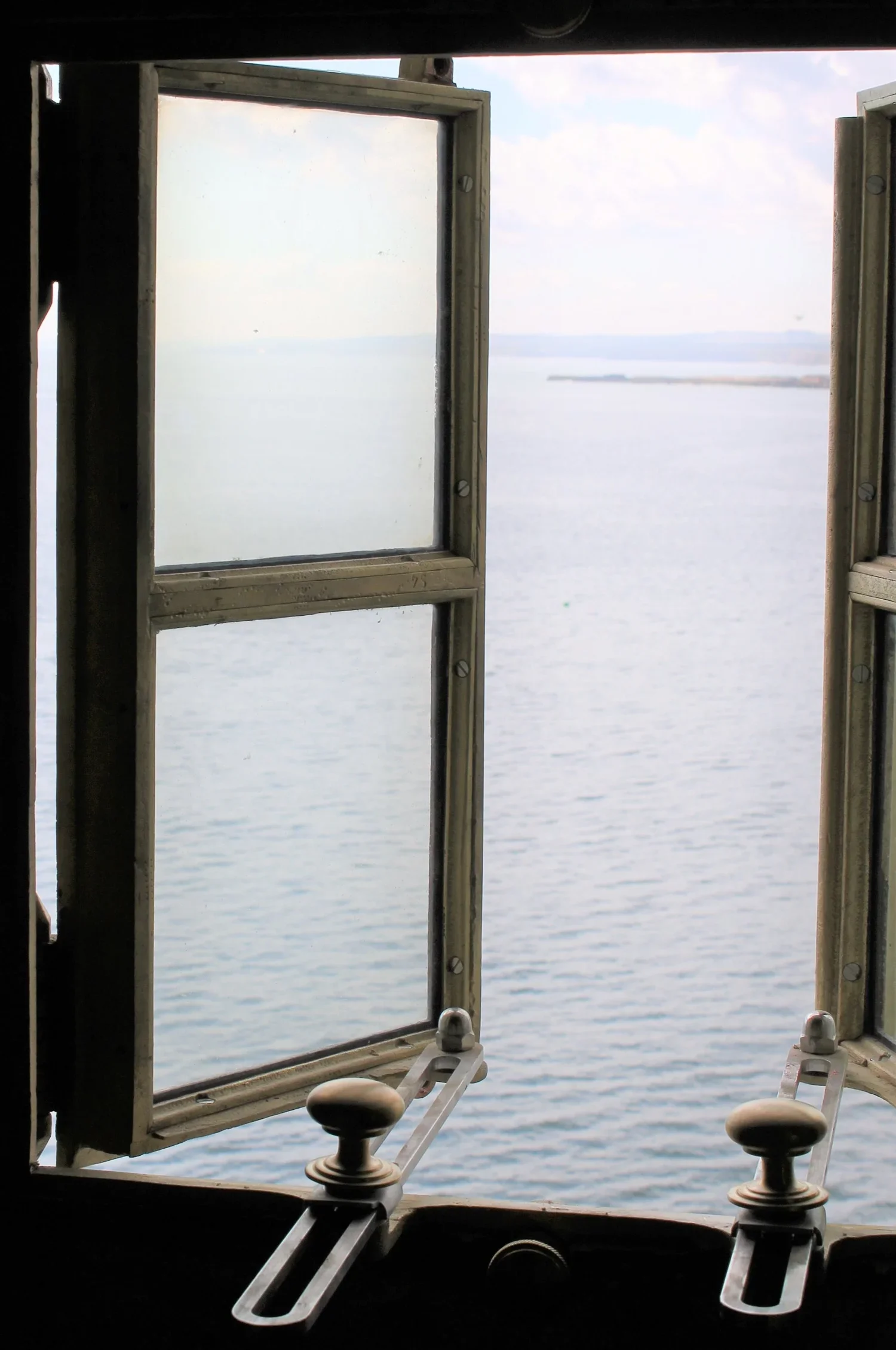 Open window overlooking a calm body of water, with a distant shoreline and cloudy sky.