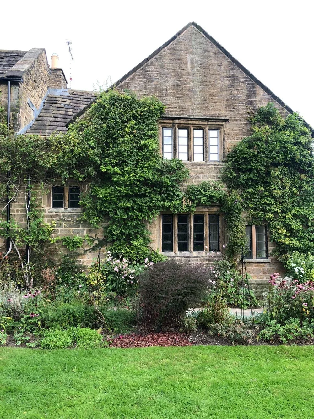 A stone house with ivy growing on the walls, surrounded by a well-kept garden with flowering plants and green grass.