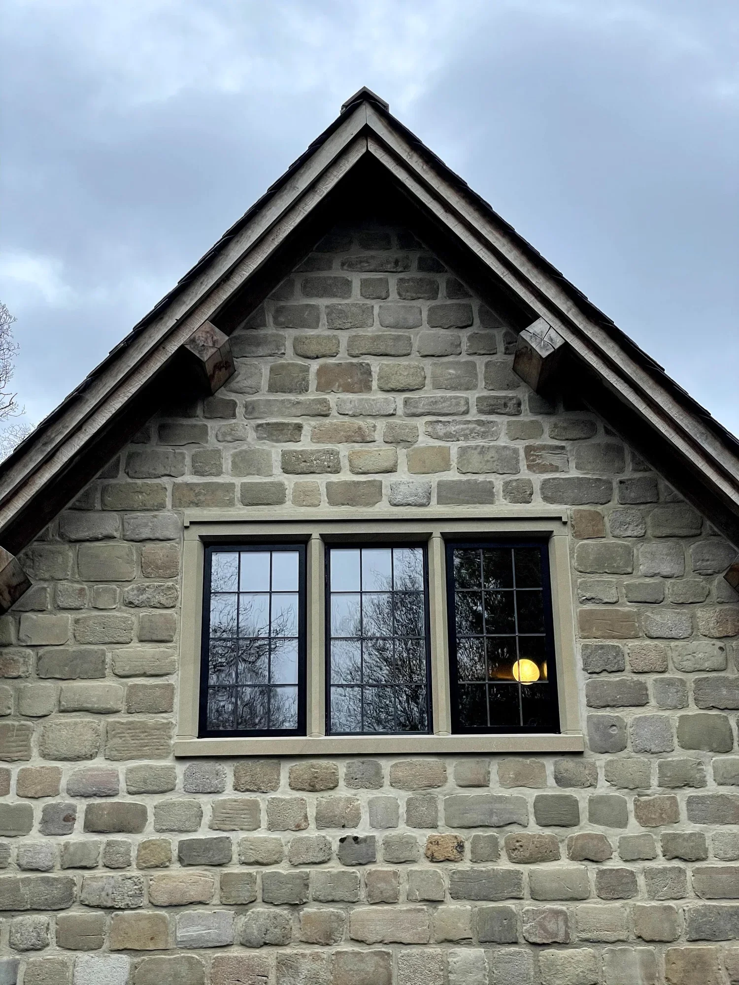 The image shows the upper part of a house with a brick wall, a pointed gable roof with shingles, and a window with divided panes reflecting trees and sky.