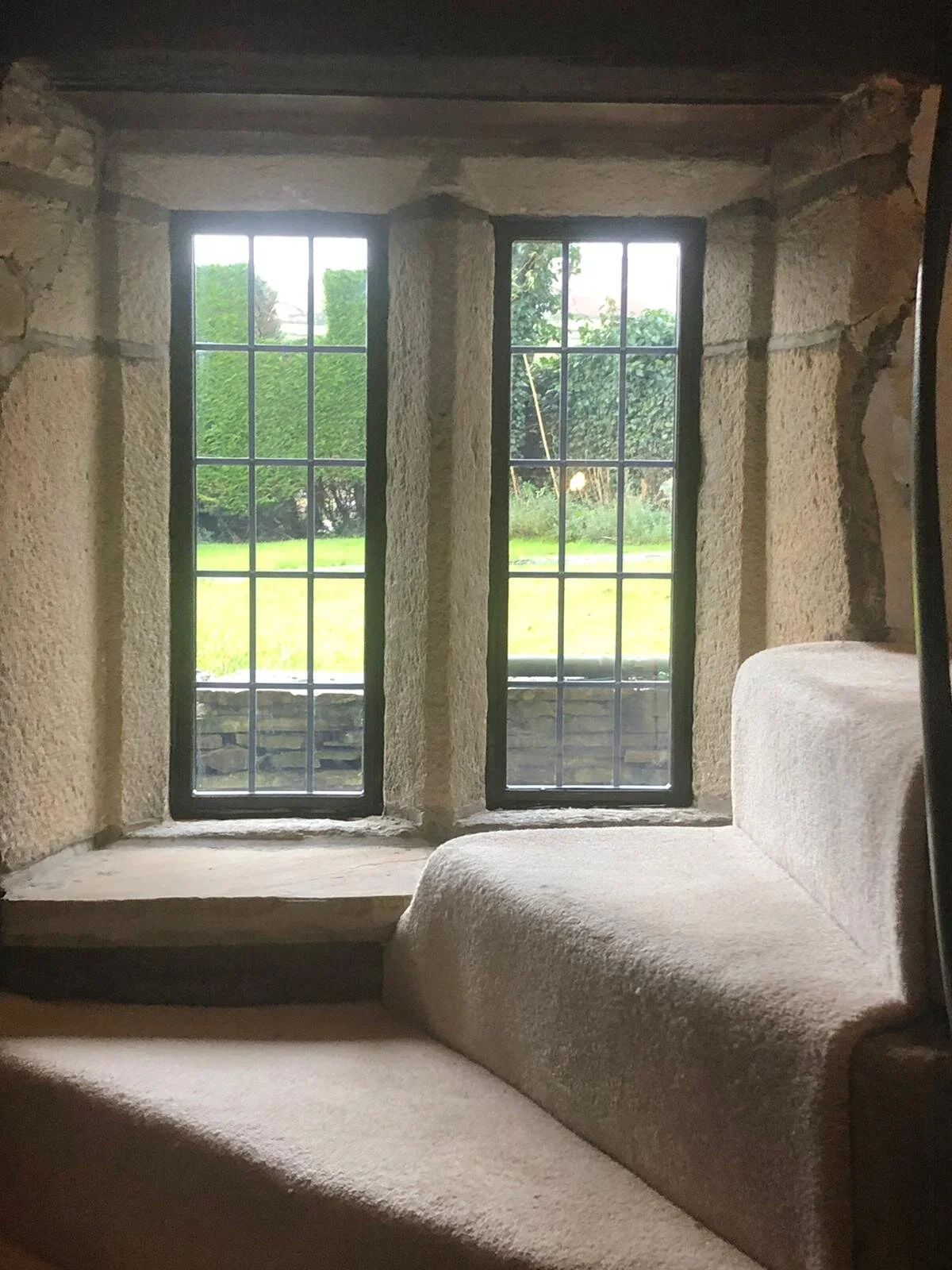 View of two large outdoor windows with metal grid panes, set in a stone wall, overlooking a garden with grass and trees, inside a room with a cushioned bench or window seat.