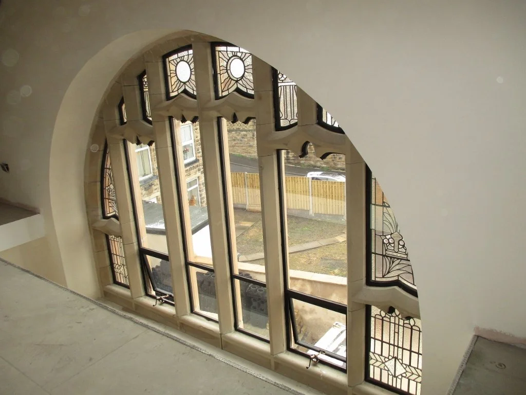 Interior view of a large arched window with decorative stained glass and leaded glass panels, looking out onto a backyard with a wooden fence and grass.