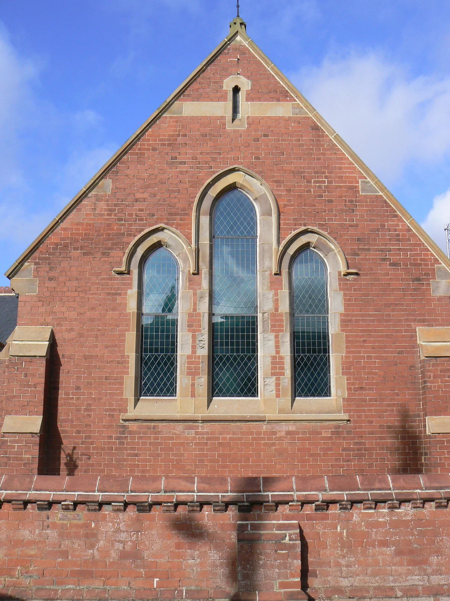 Front view of a red brick church facade with three tall, narrow arched windows with diamond-patterned glass, a gable roof, and a small narrow window at the top under the roof peak, against a background of a blue sky with some clouds.